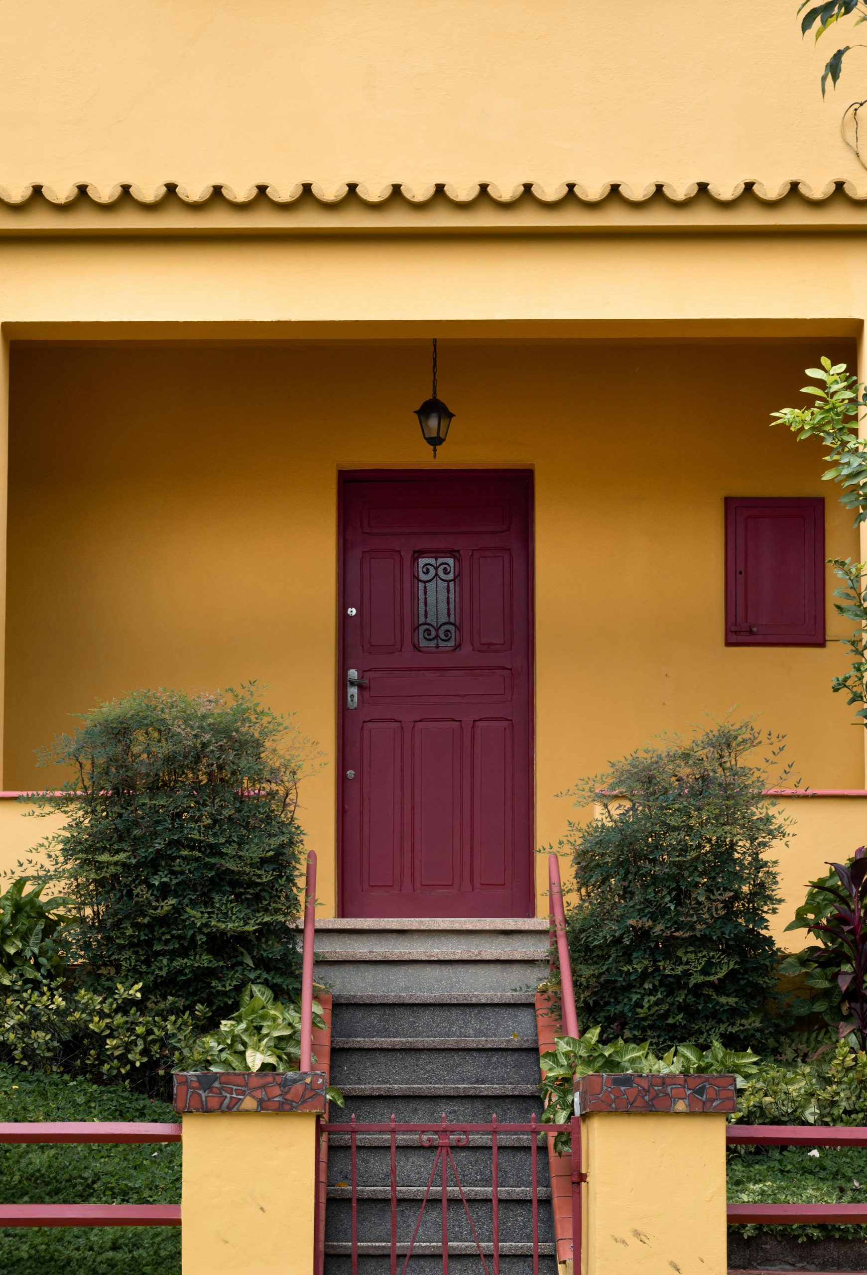 Charming yellow house facade with a maroon door and green shrubs accentuating the entrance.