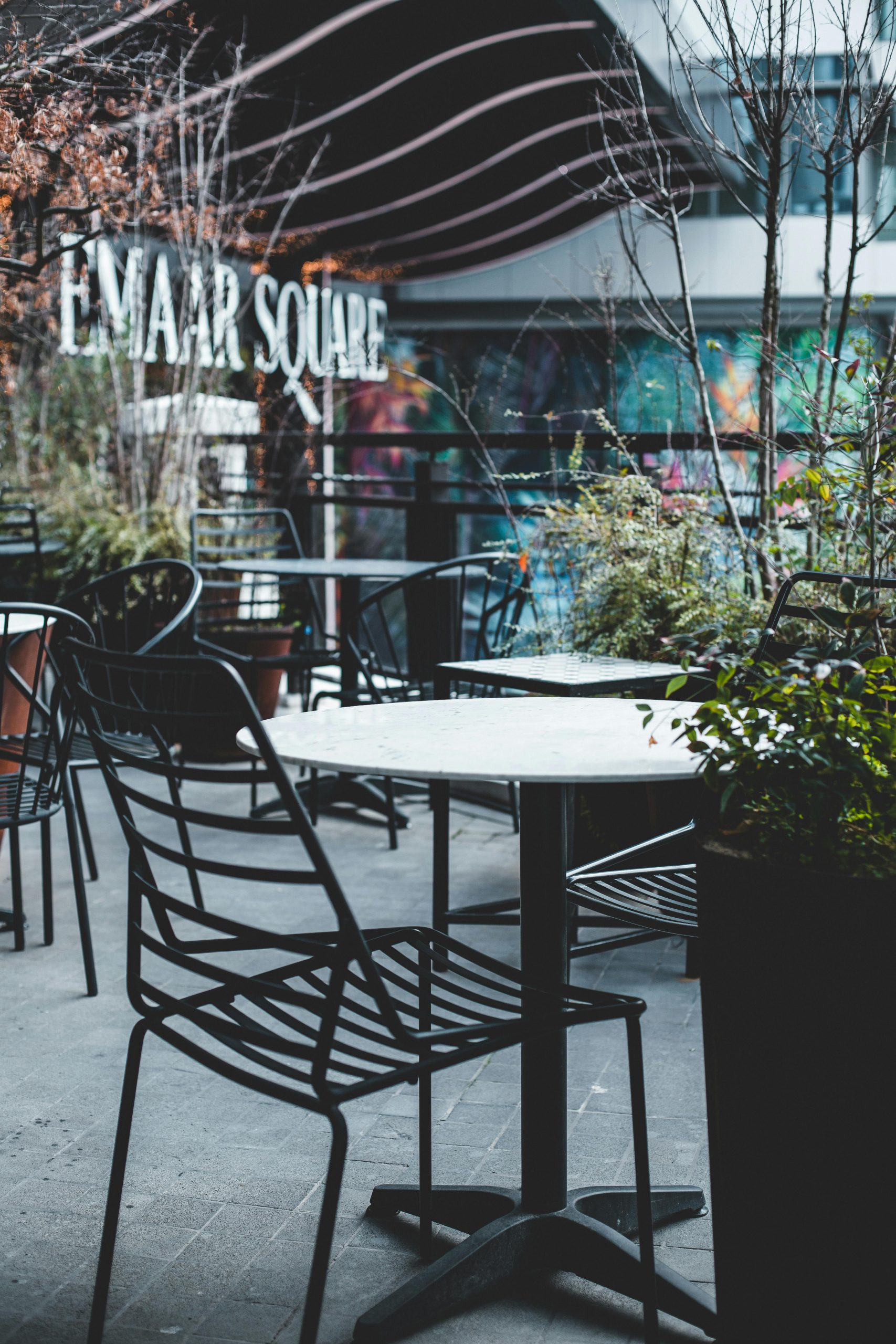 Empty terrace at a modern café, Emaar Square, with metal chairs, tables, and potted plants.
