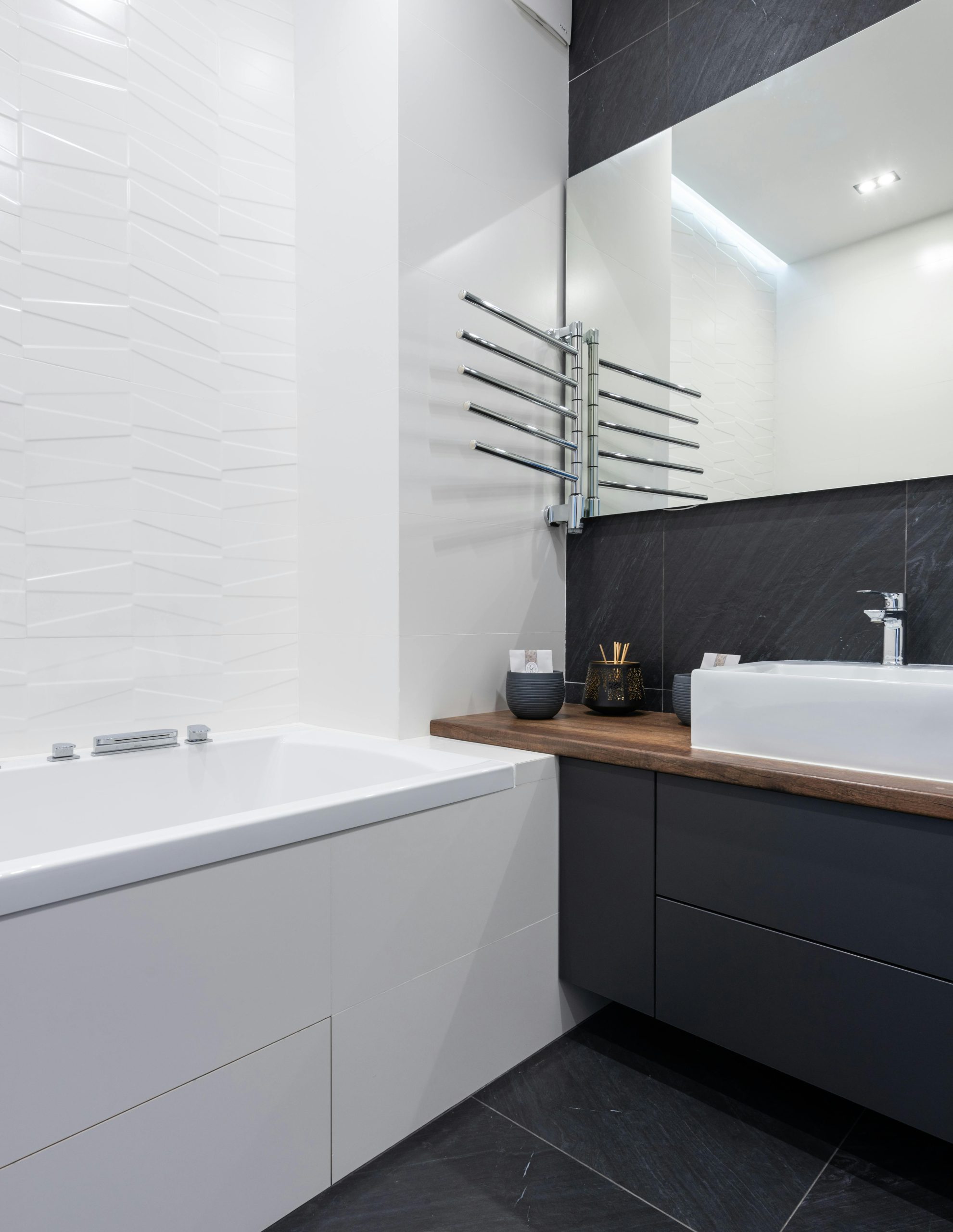 Interior of modern washroom with mirror near white ceramic bath placed near black cupboard with sink and toiletry at home