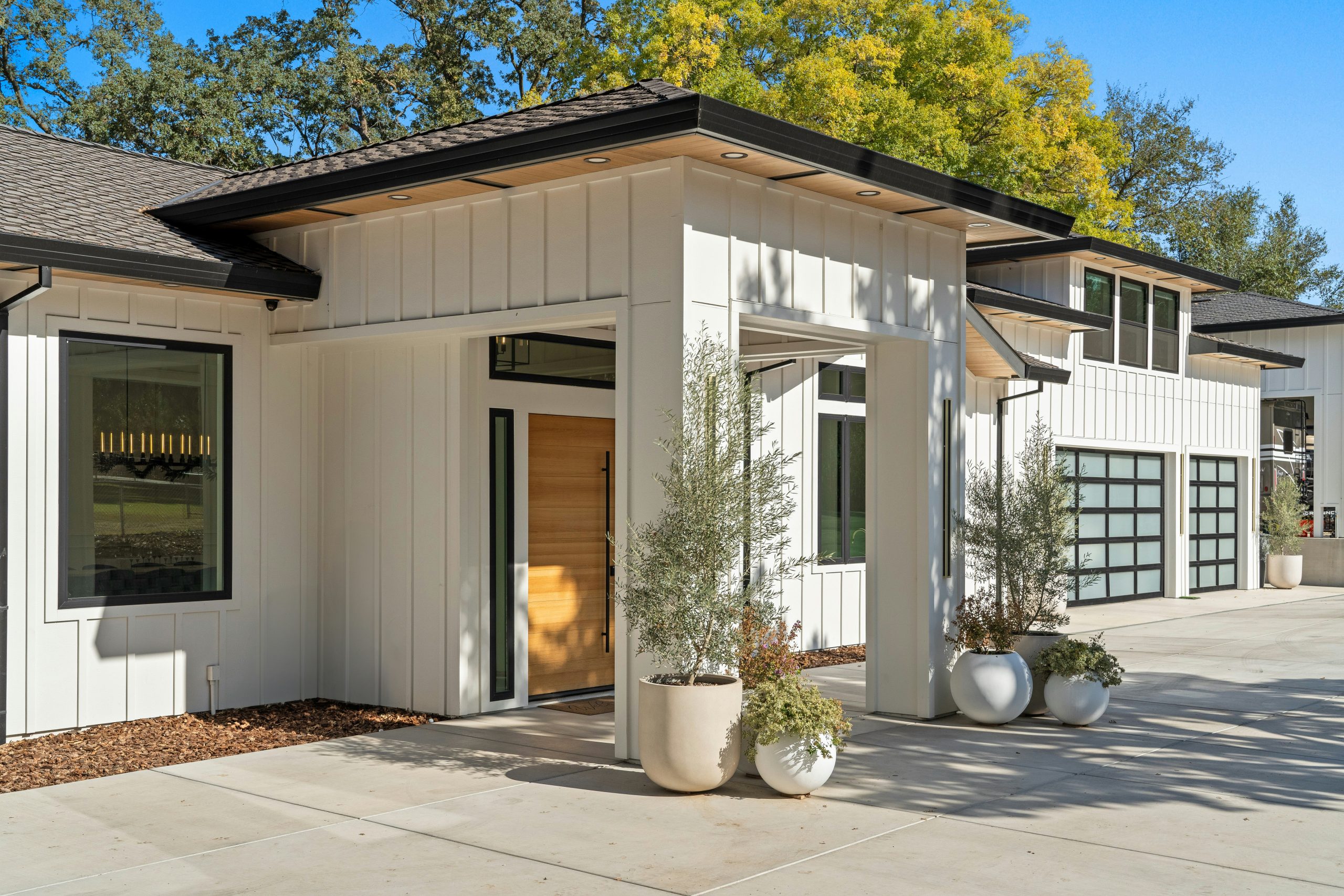 Modern farmhouse exterior with white board and batten siding, black trim, and natural wood front door. Luxury custom home with large windows, contemporary garage doors, and clean landscaping under bri