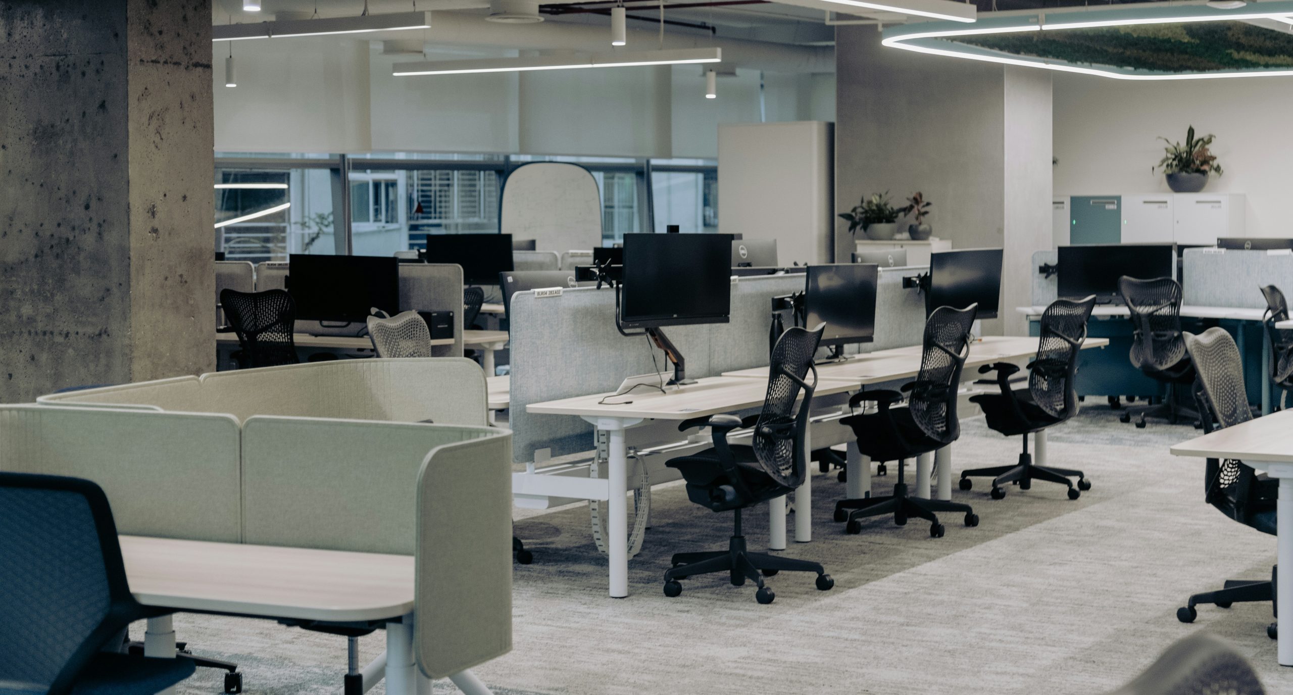 The image depicts a modern, open-plan office with a clean and organized layout. The workspace features multiple desks equipped with computers and ergonomic chairs. The desks are separated by low divid