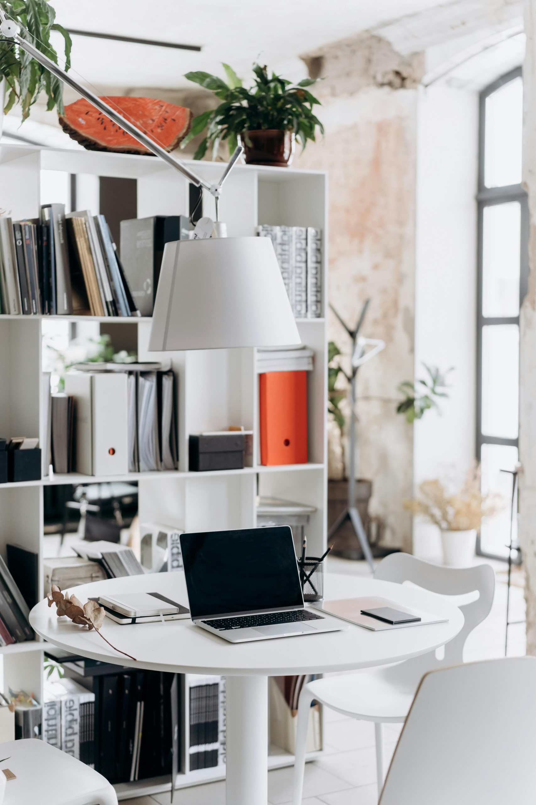 Well-lit modern office with a laptop on a round table, surrounded by shelves and plants.