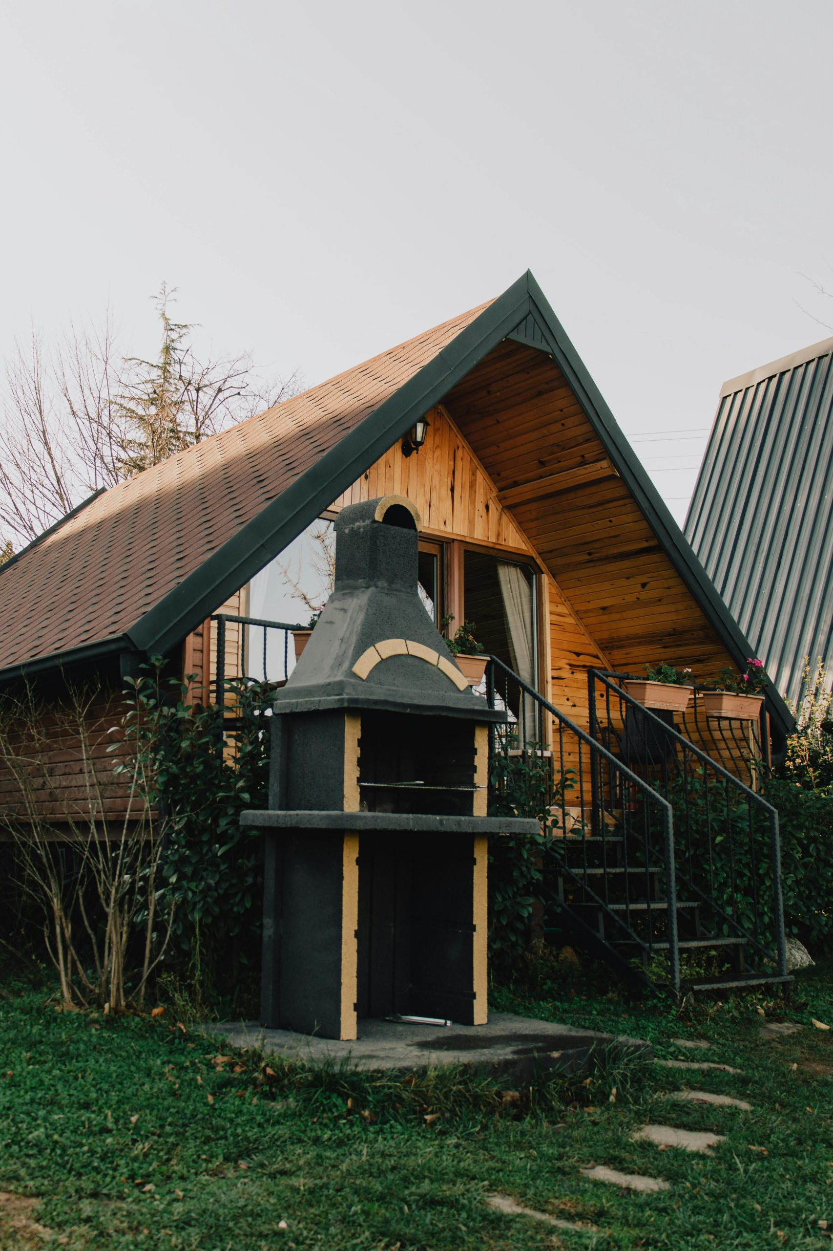 A cozy wooden cabin featuring a triangular roof and outdoor stone fireplace surrounded by greenery.