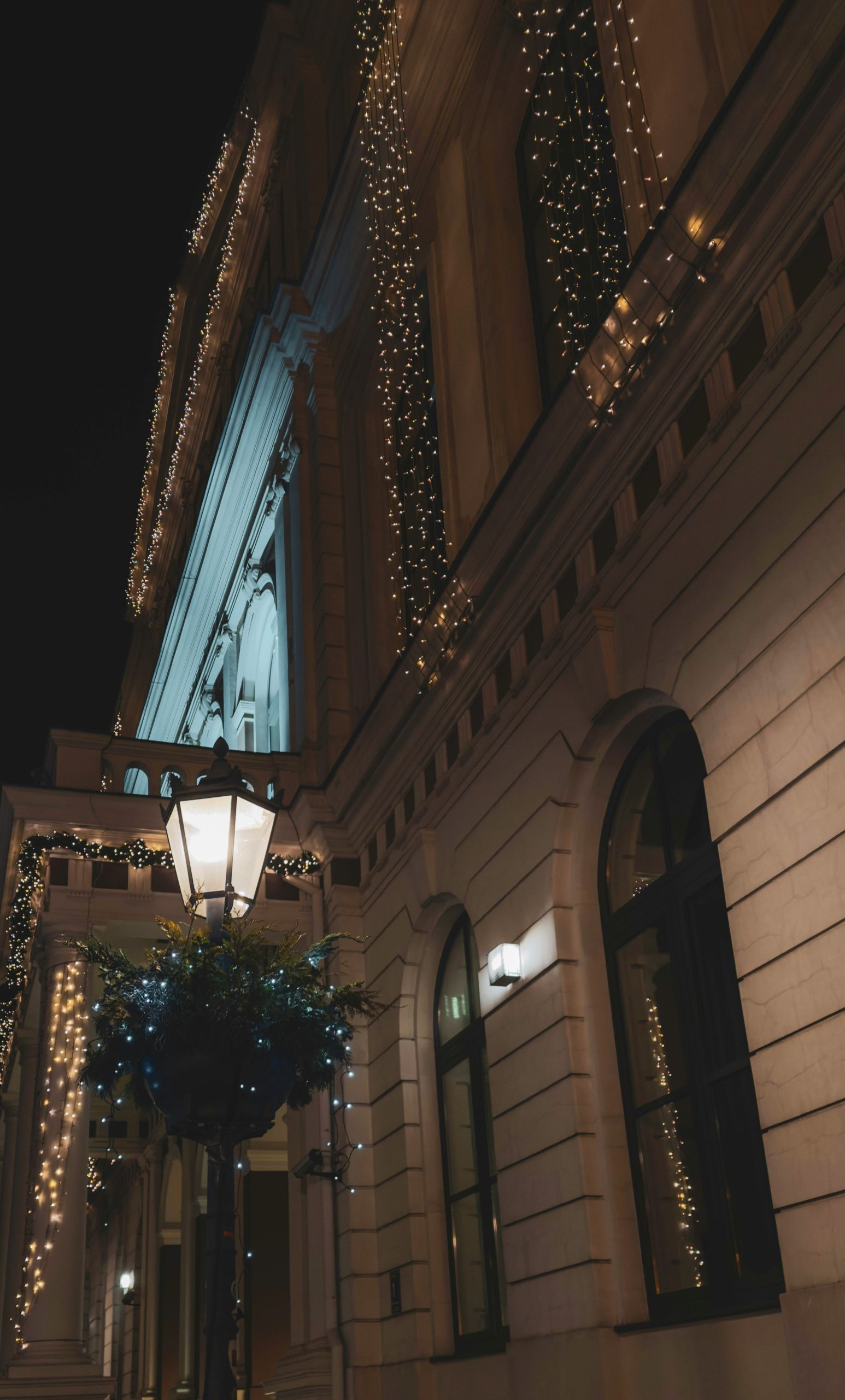 An illuminated, decorated street lamp in front of a building adorned with festive Christmas lights at night