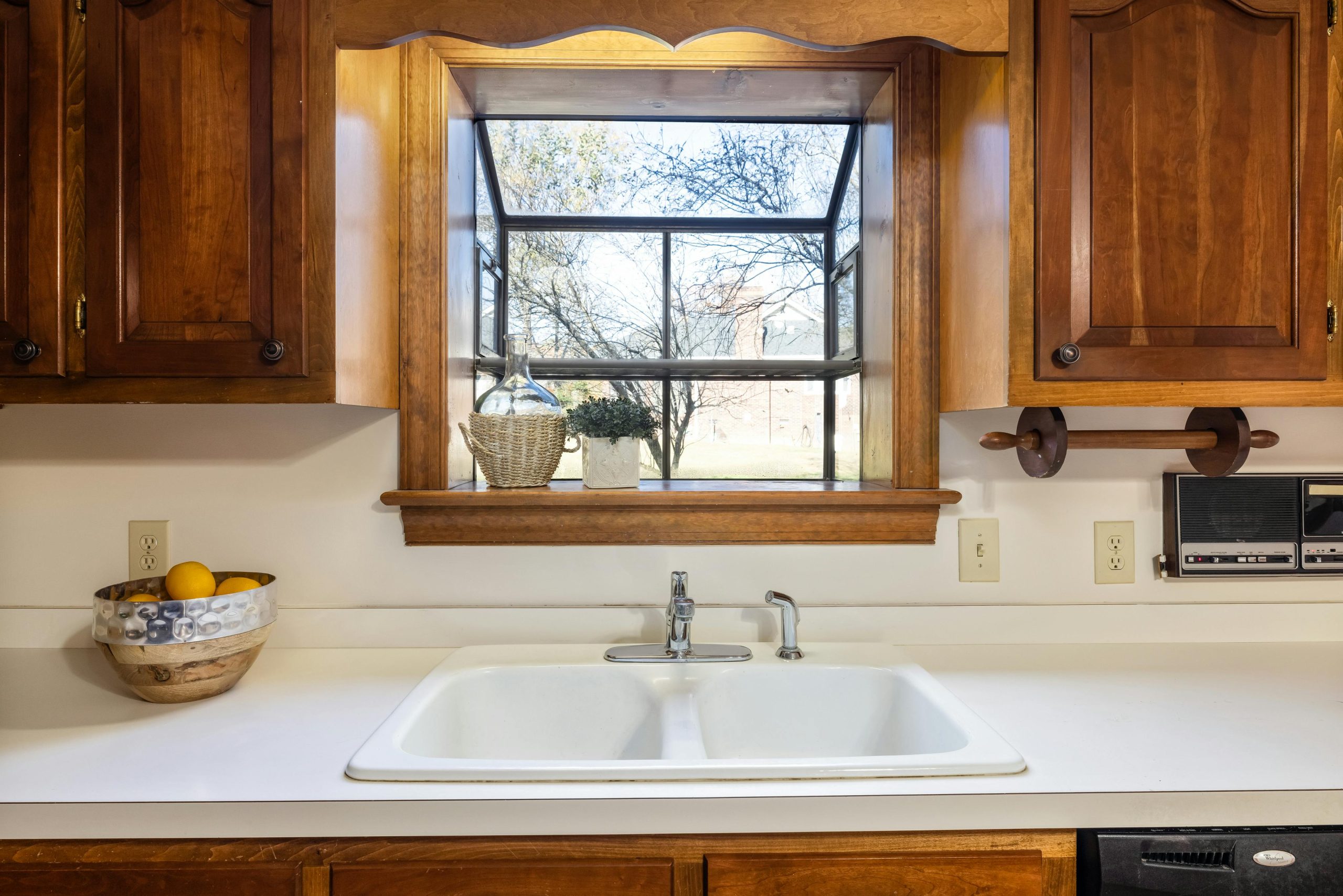 Bright kitchen interior featuring a ceramic sink, wooden cabinets, and a view through a large window.