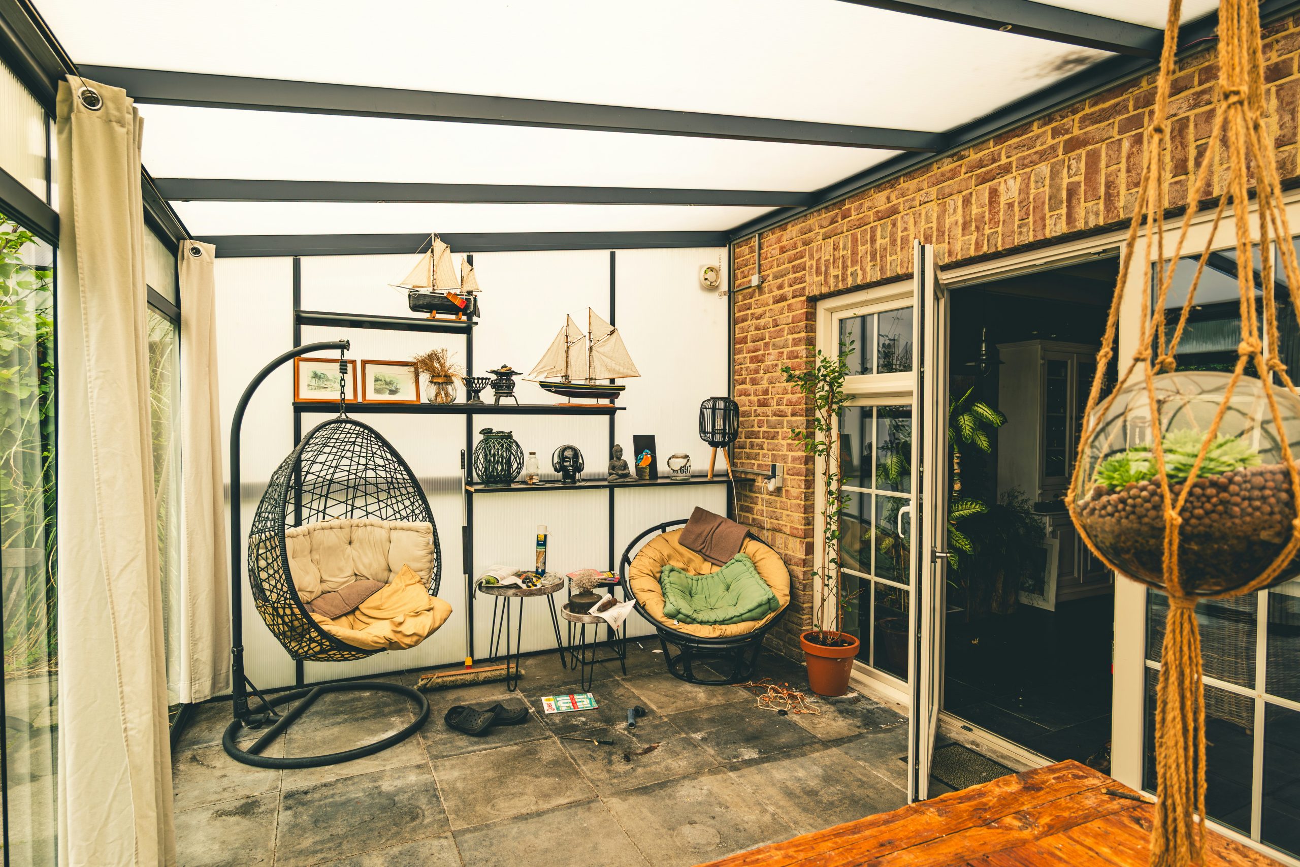 Covered terrace of a guesthouse in Schoonoord, Netherlands, furnished with hanging chairs, lounge seating, and decorative objects. This semi-outdoor space functions as a relaxation area, bridging inte