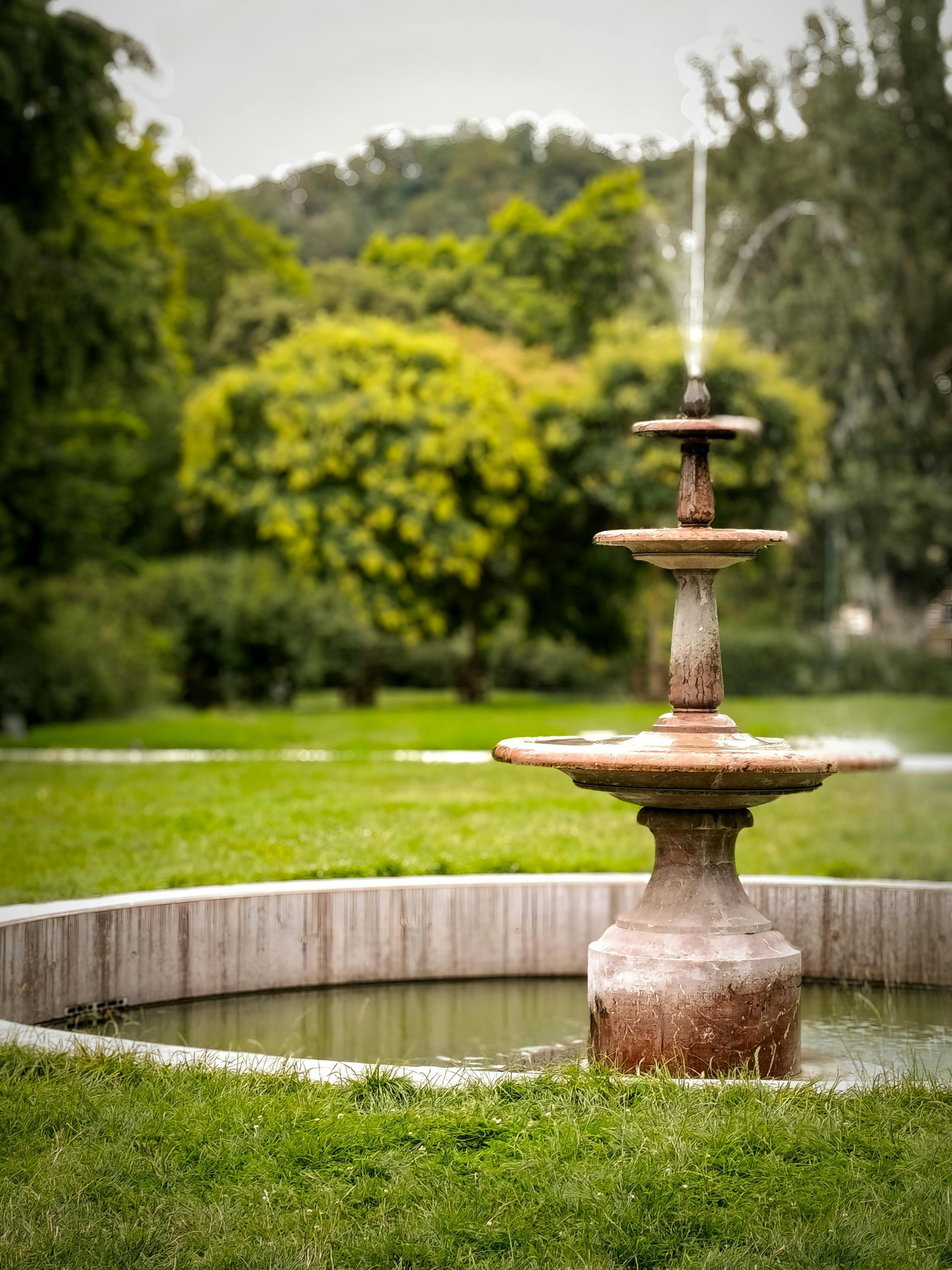 Old stone fountain in a serene park setting, surrounded by vivid greenery, creating a tranquil atmosphere.