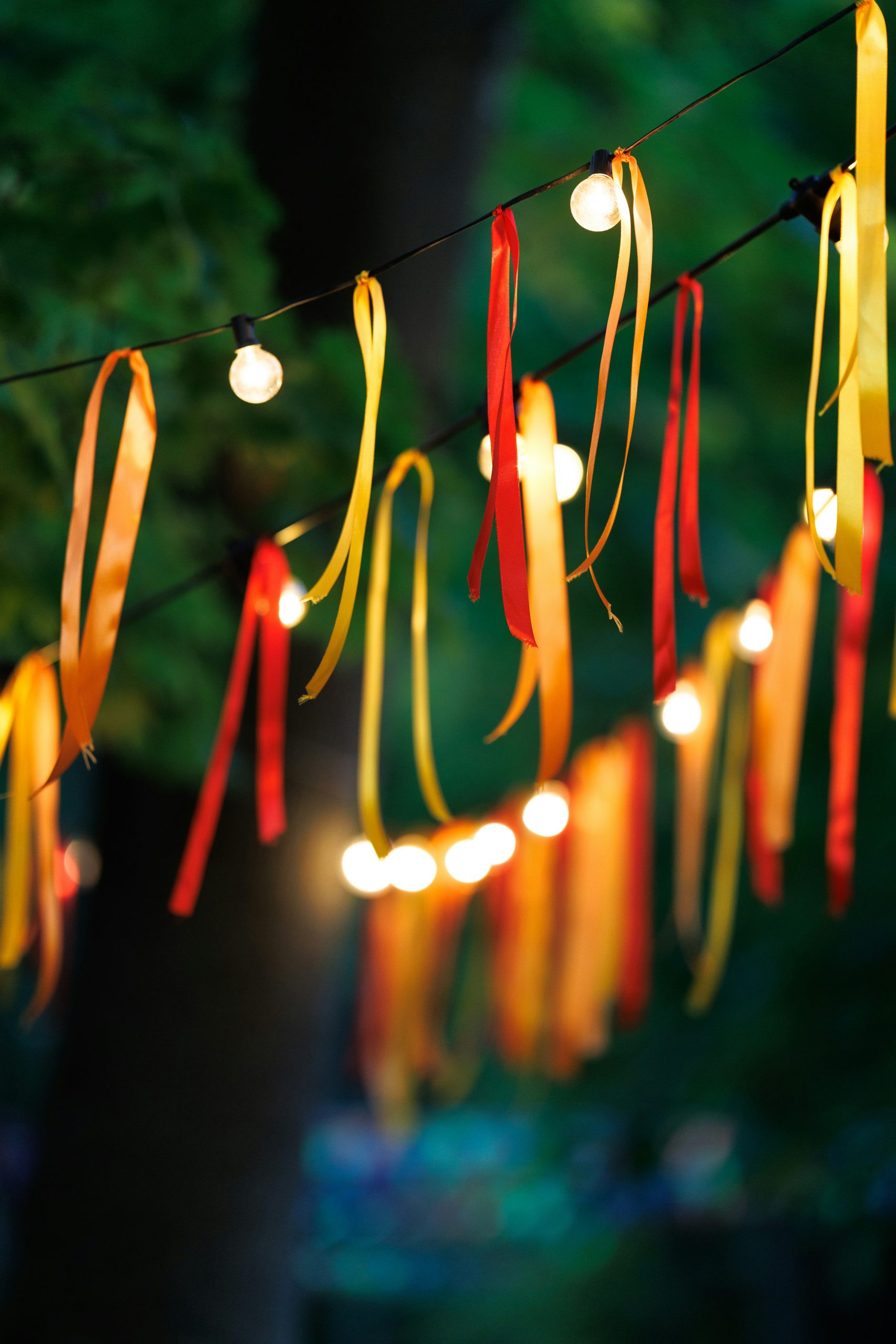 String lights adorned with flowing red and yellow ribbons hang outdoors, creating a festive and cheerful atmosphere. The bright colors and soft illumination evoke celebration, warmth, and a playful to