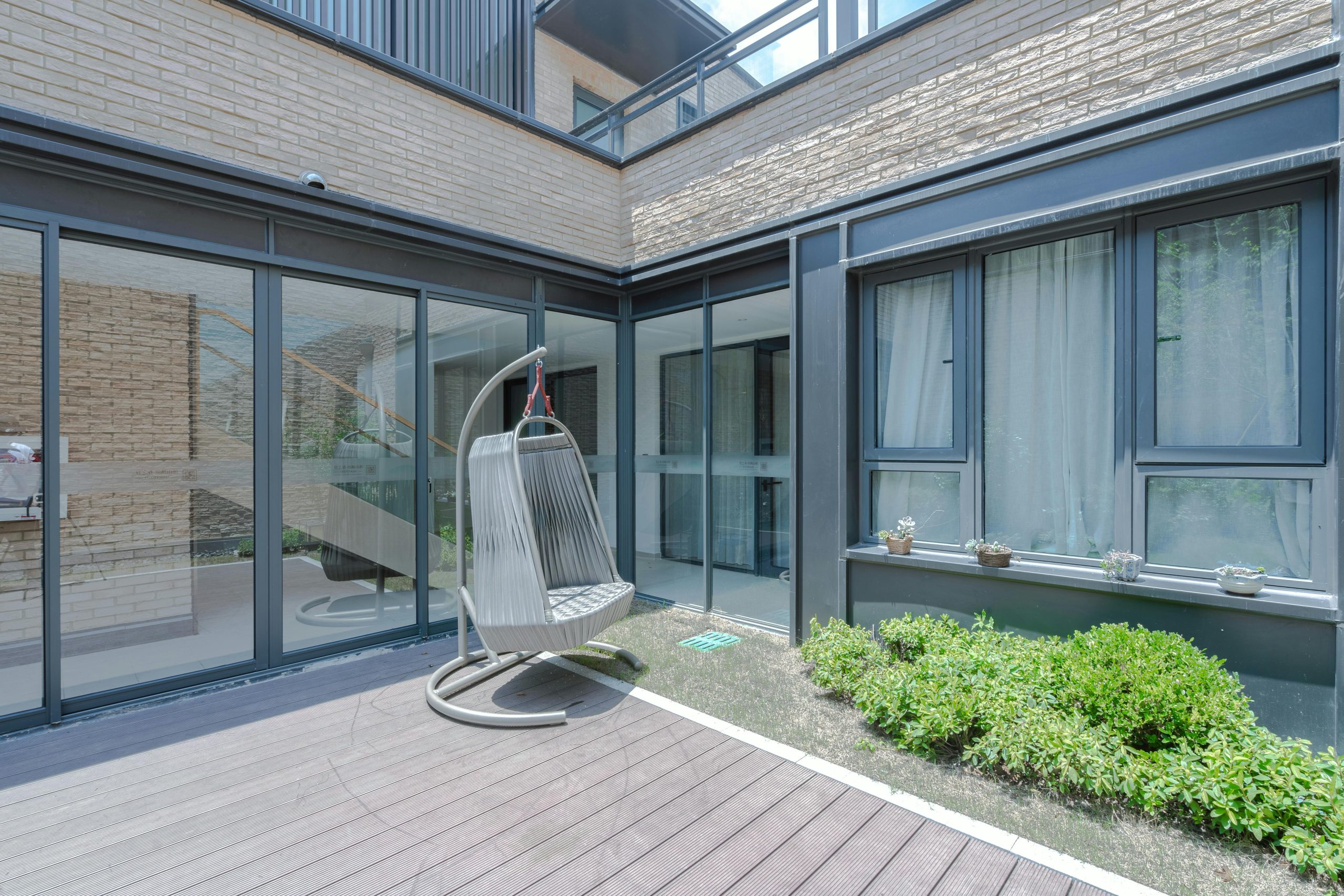 Sunny modern patio featuring elegant glass walls, a cozy rattan swing, and lush greenery.