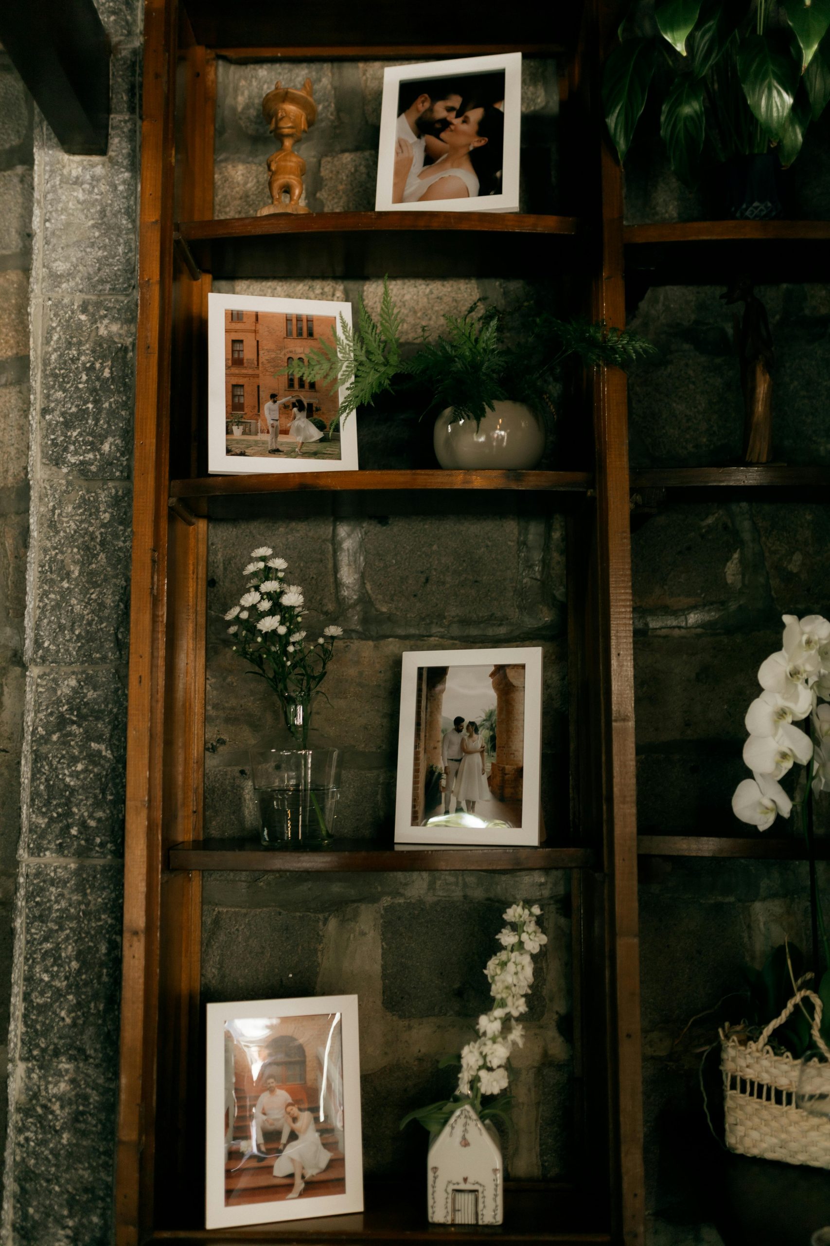 Wooden shelf featuring framed photos, plants, and flowers on a stone wall.