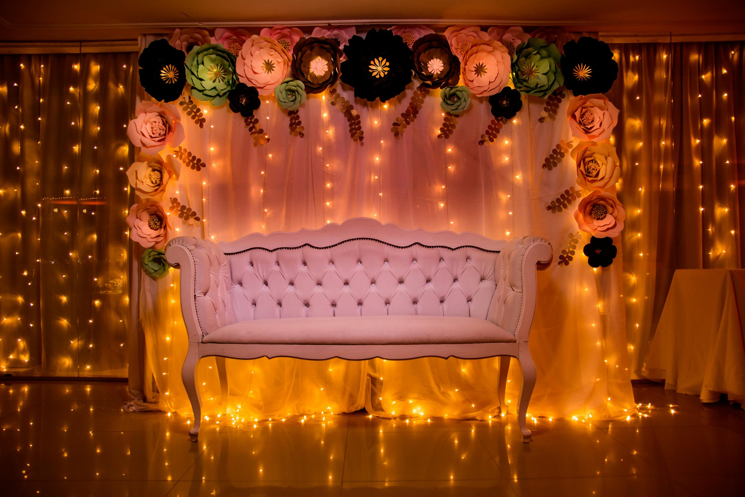 A beautifully decorated photo backdrop with a white sofa, paper flowers, and warm fairy lights.