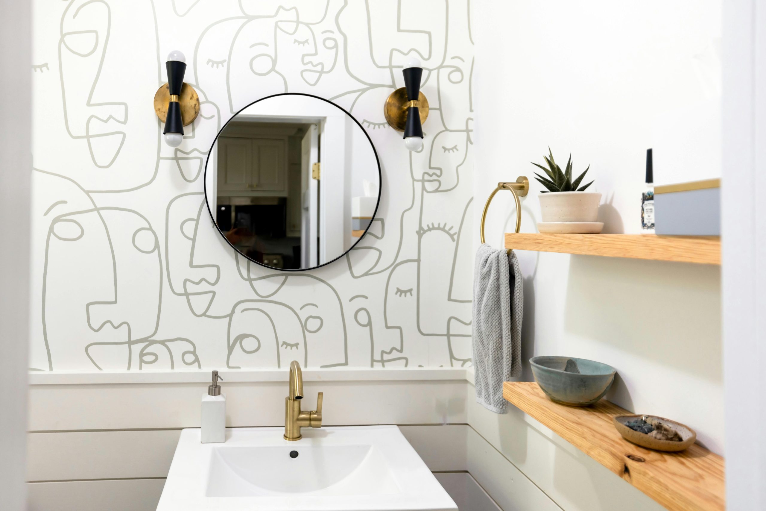A contemporary bathroom featuring abstract line art wallpaper, round mirror, and minimalist wooden shelves.