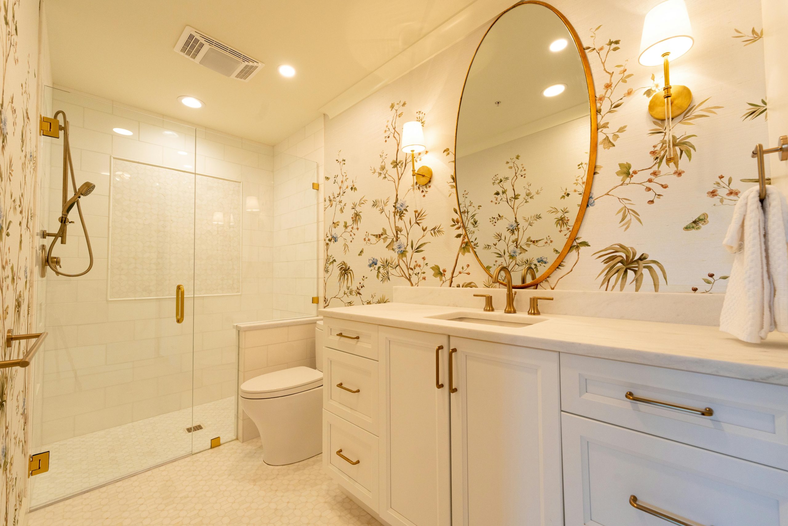 A modern bathroom with elegant floral wallpaper, gold fixtures, and a large round mirror.