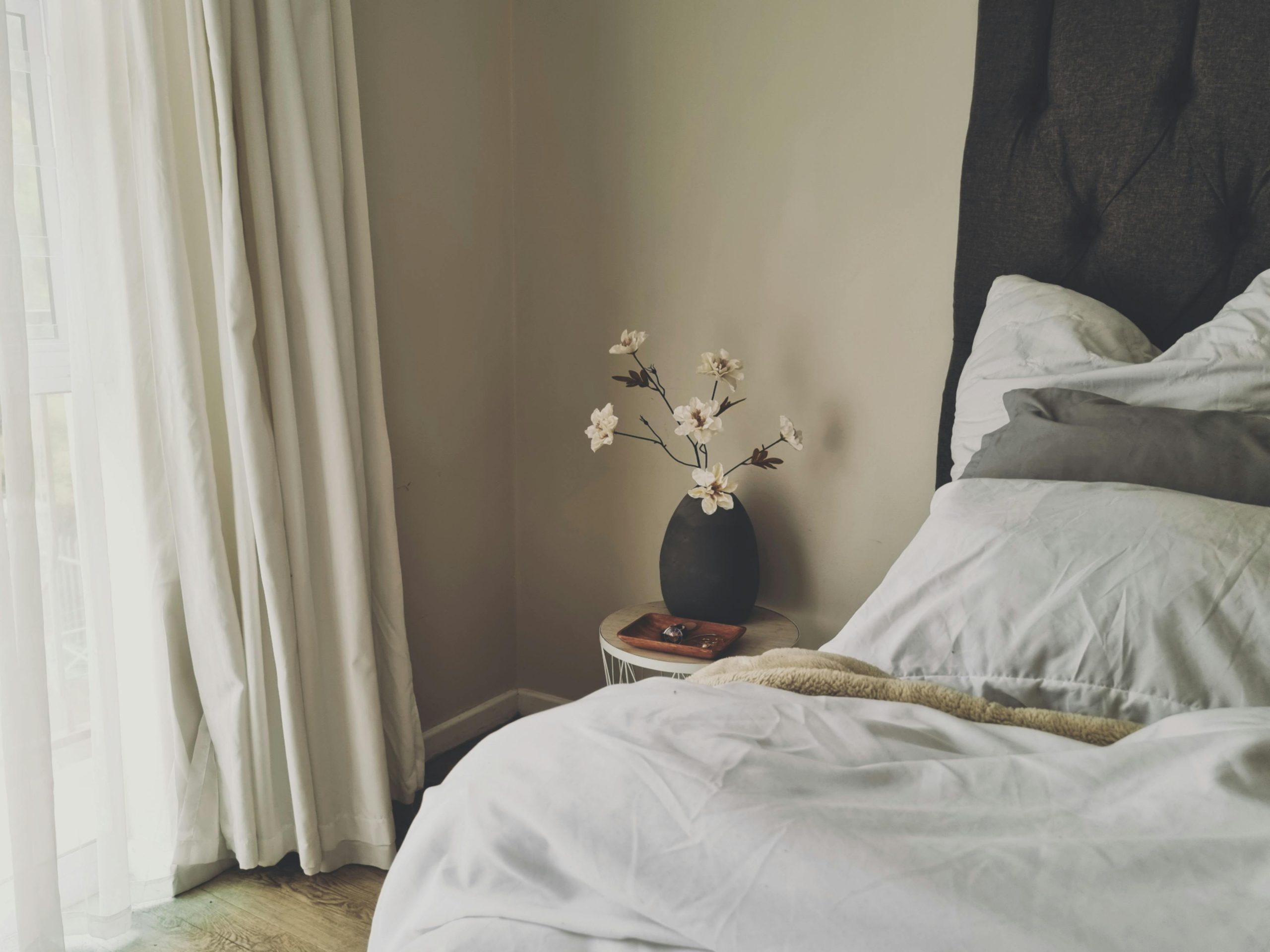 A serene and minimalist bedroom interior featuring white flowers in a vase, natural light, and a cozy bedding setup.