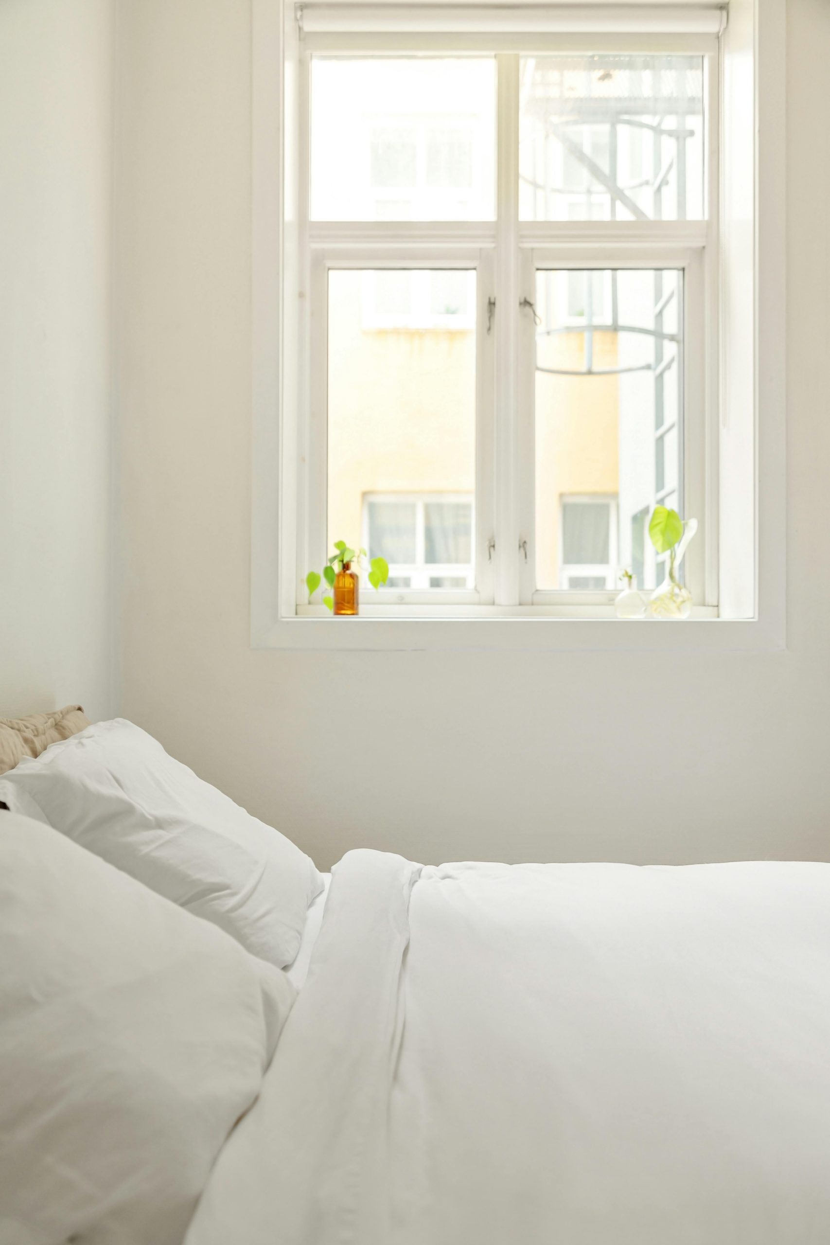 A serene minimalist bedroom featuring white beddings and natural light through a window.