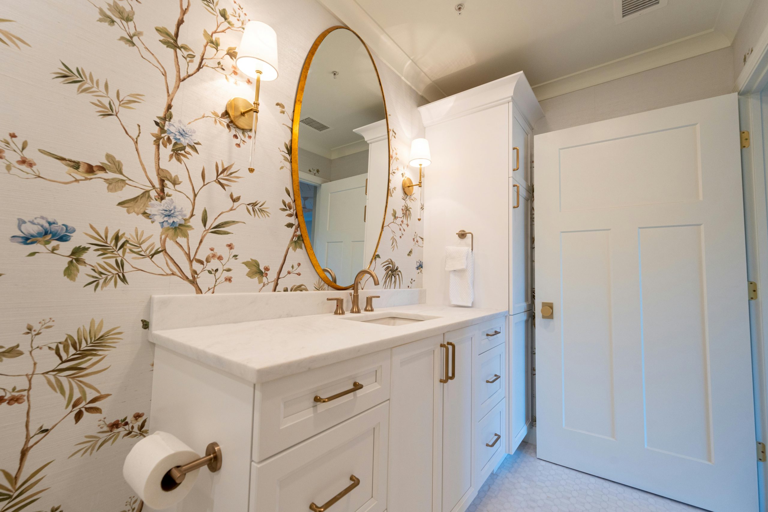 A stylish bathroom interior featuring floral wallpaper, white cabinetry, and gold accents.