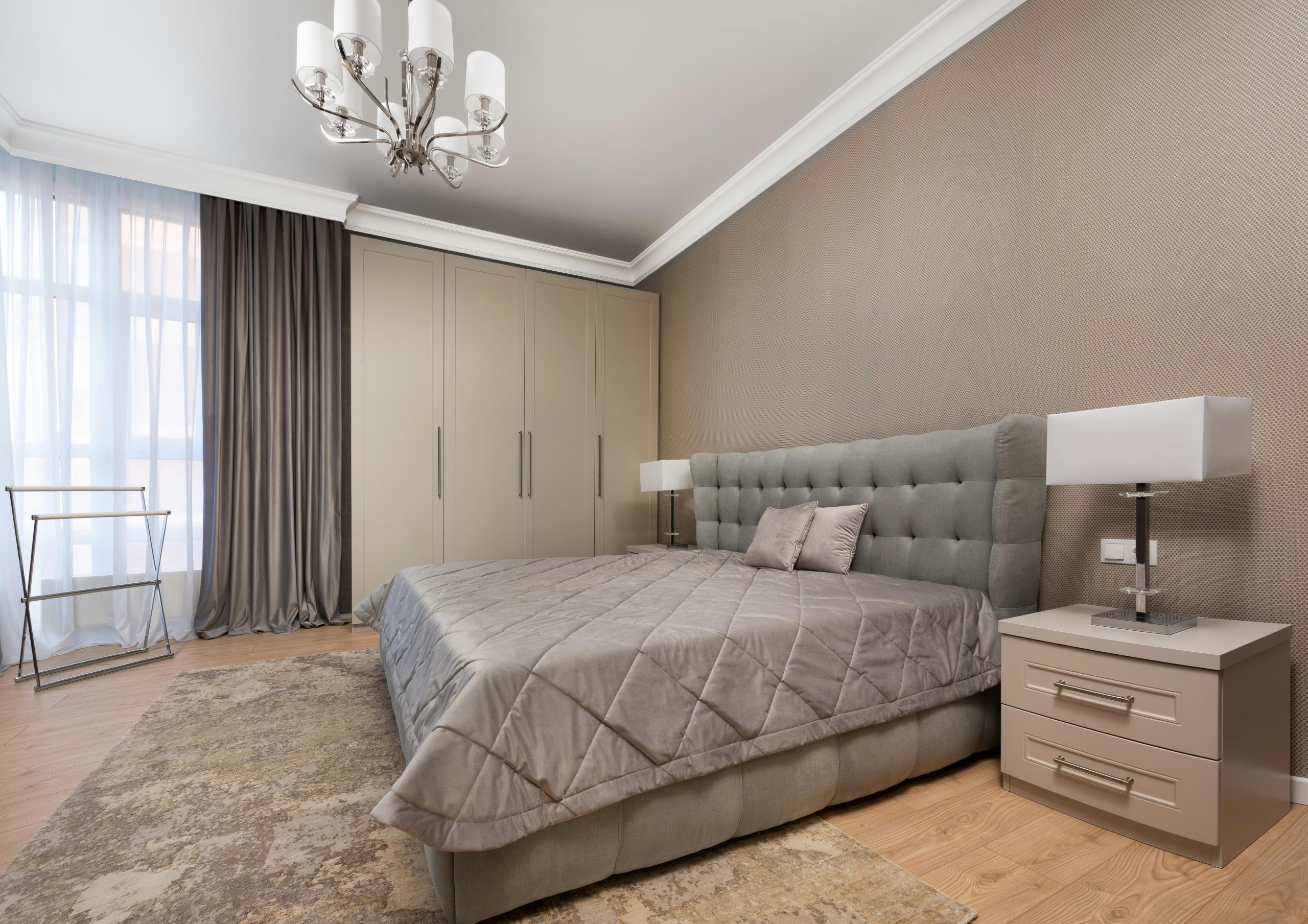 Bed with coverlet and cushions between bedside tables with lamps in modern bedroom with chandelier in daylight