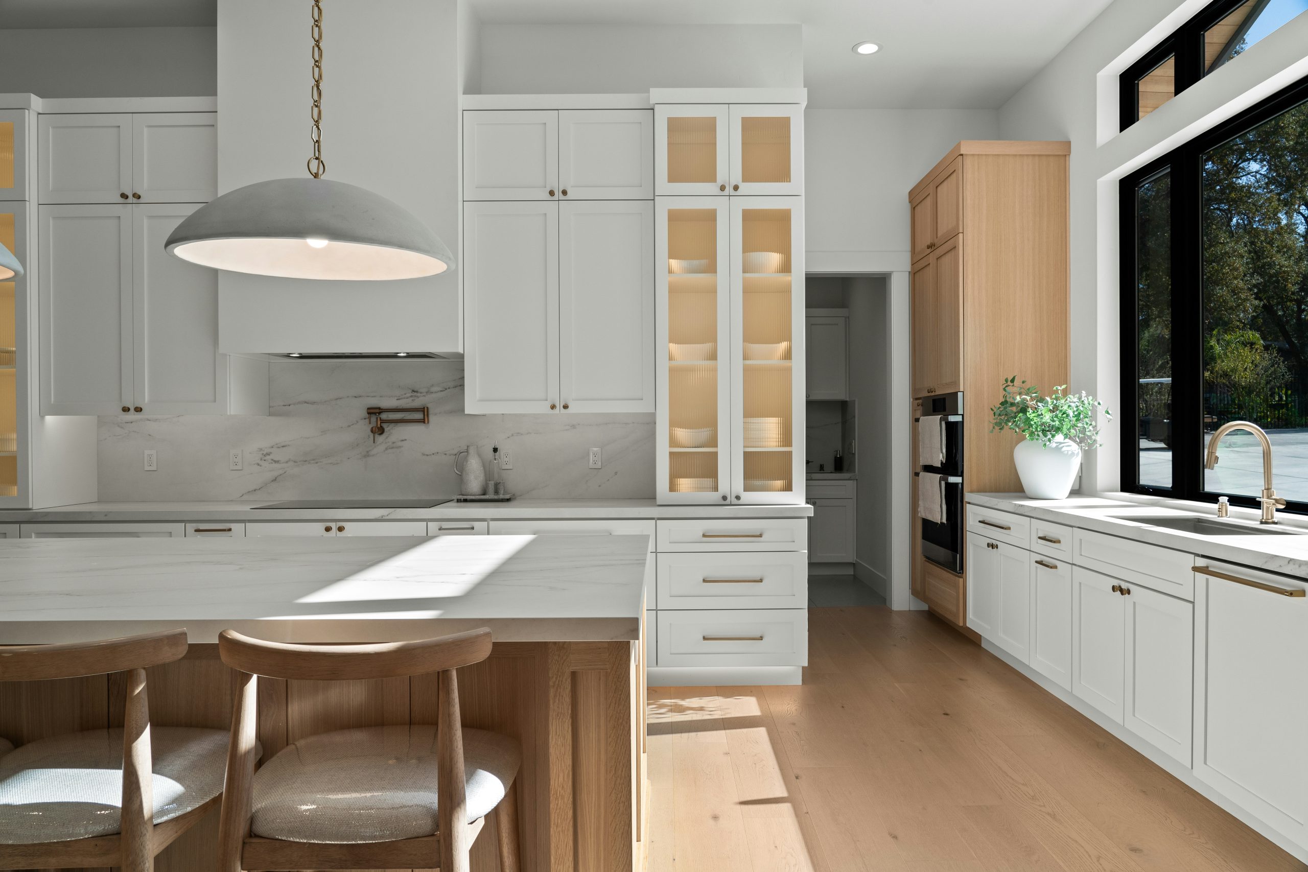 Bright modern kitchen with white shaker cabinets, warm wood accents, and marble backsplash. Contemporary custom home featuring a large island, brass hardware, statement pendant lighting, and black-fra