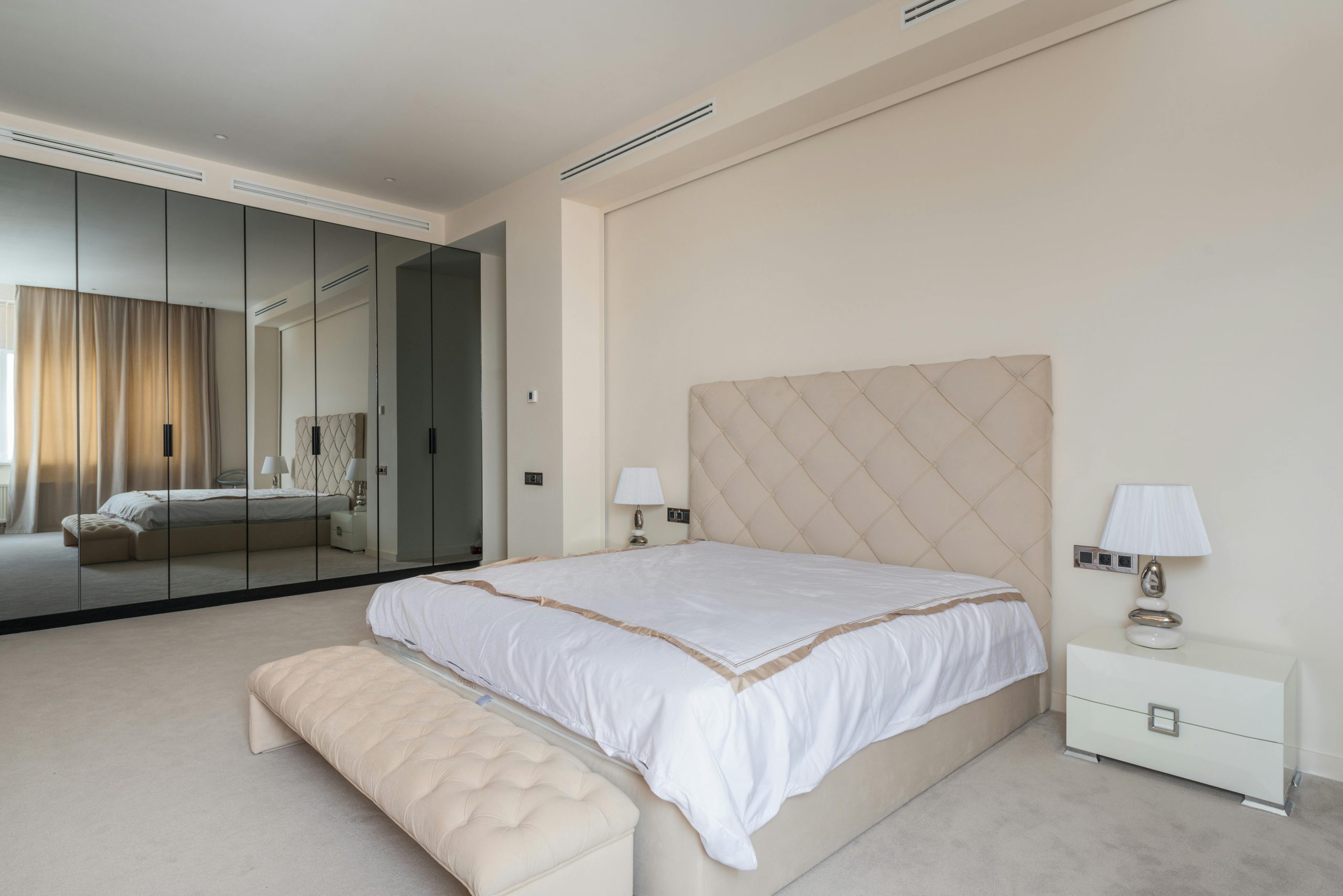 Comfortable be with beige soft headboard and ottoman bench reflecting in mirrored doors of large wardrobe in spacious classic styled bedroom in daylight
