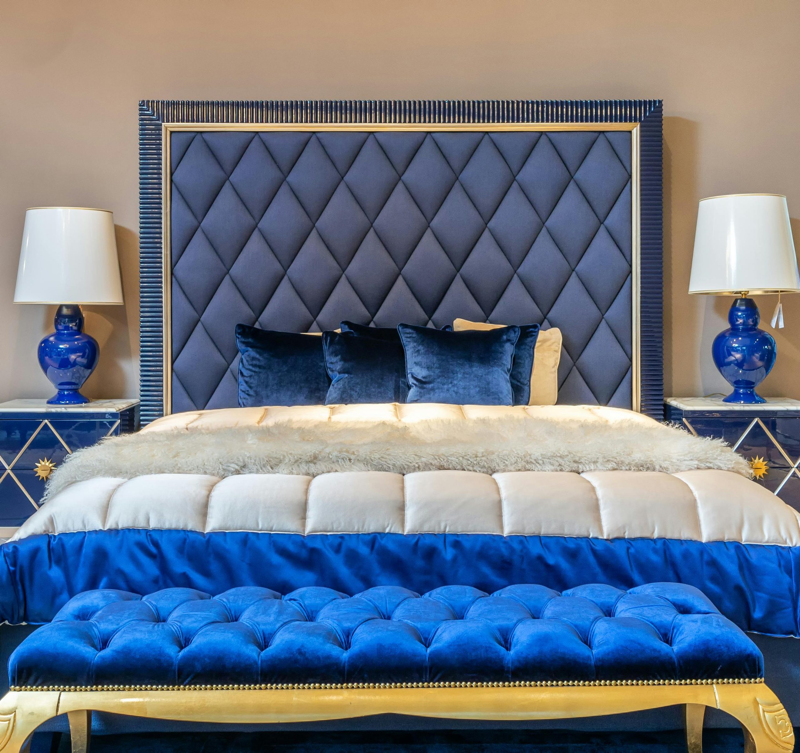 Comfortable bed with cushions and blanket near upholstered bench placed at wall between bedside tables with lamps in spacious bedroom