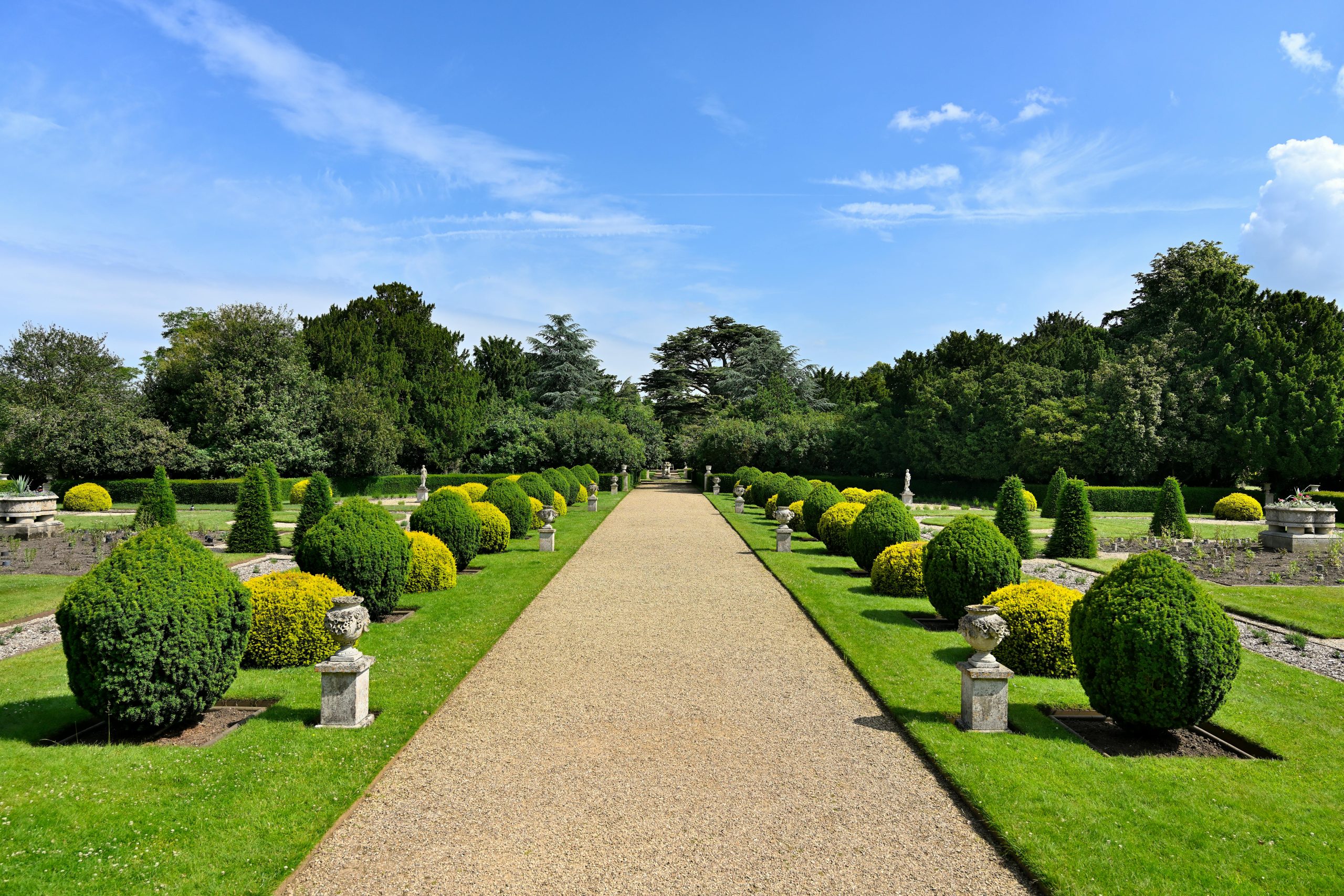 Elegant garden pathway lined with manicured bushes and classic pedestals under a clear sky.