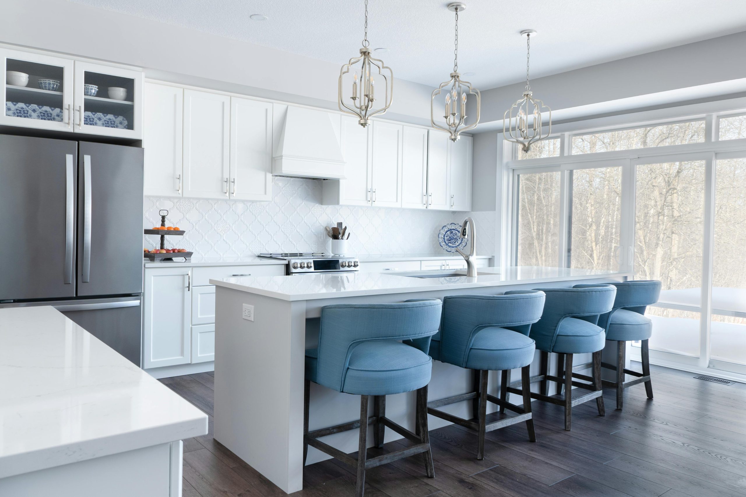 Elegant modern kitchen featuring white cabinets, blue bar stools, and large windows overlooking a snowy outdoors.