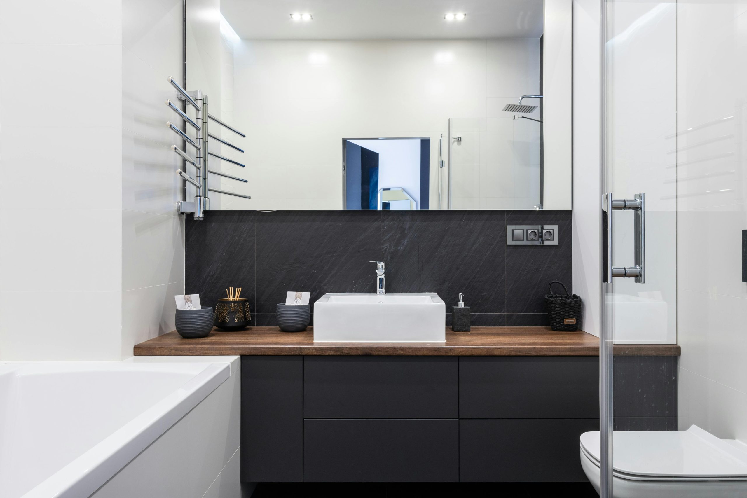 Interior of contemporary bathroom with shower cabin white tub and toilet near black cupboard with sink placed in room with mirror