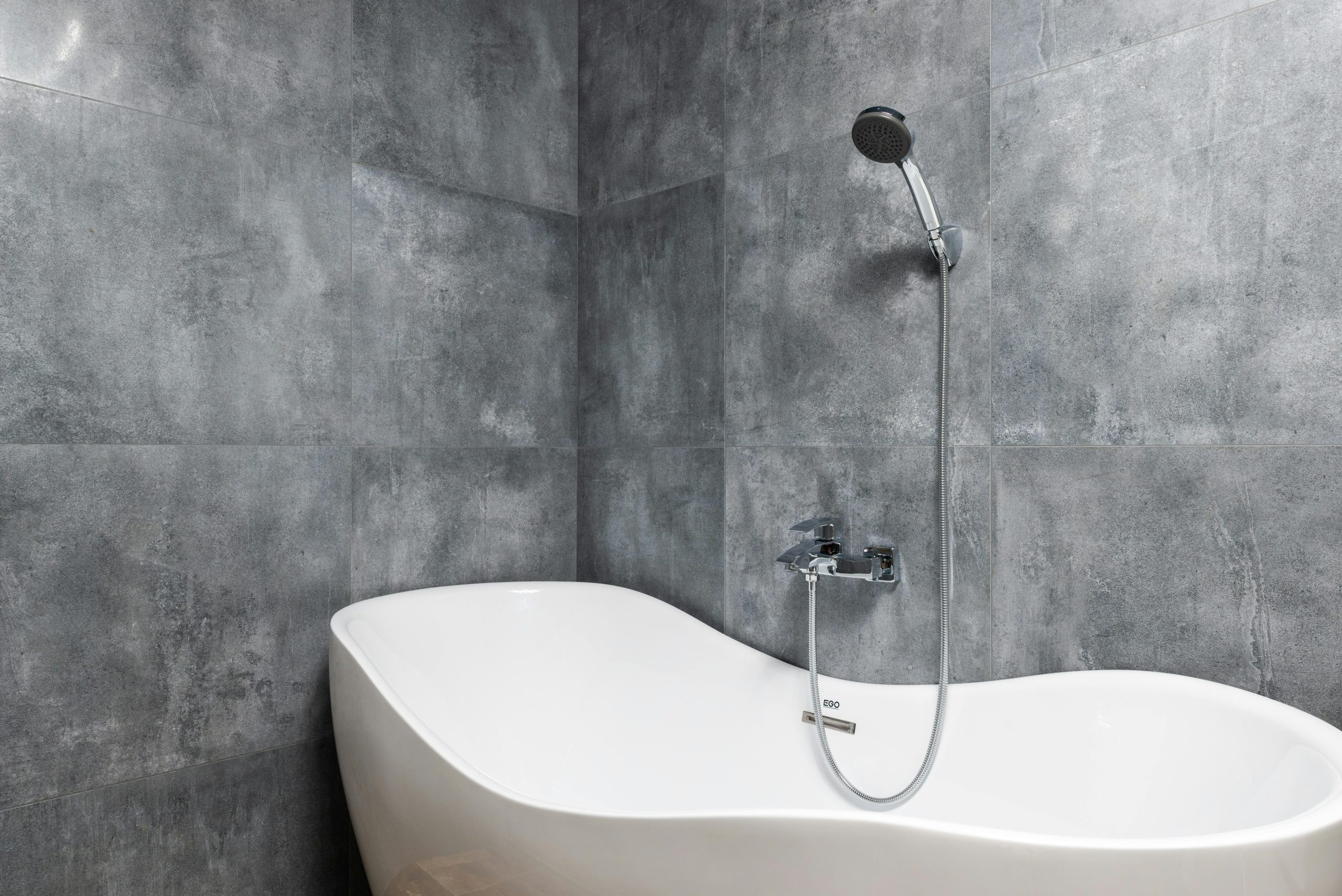 Interior of light modern bathroom with white bath under shower on gray tile wall