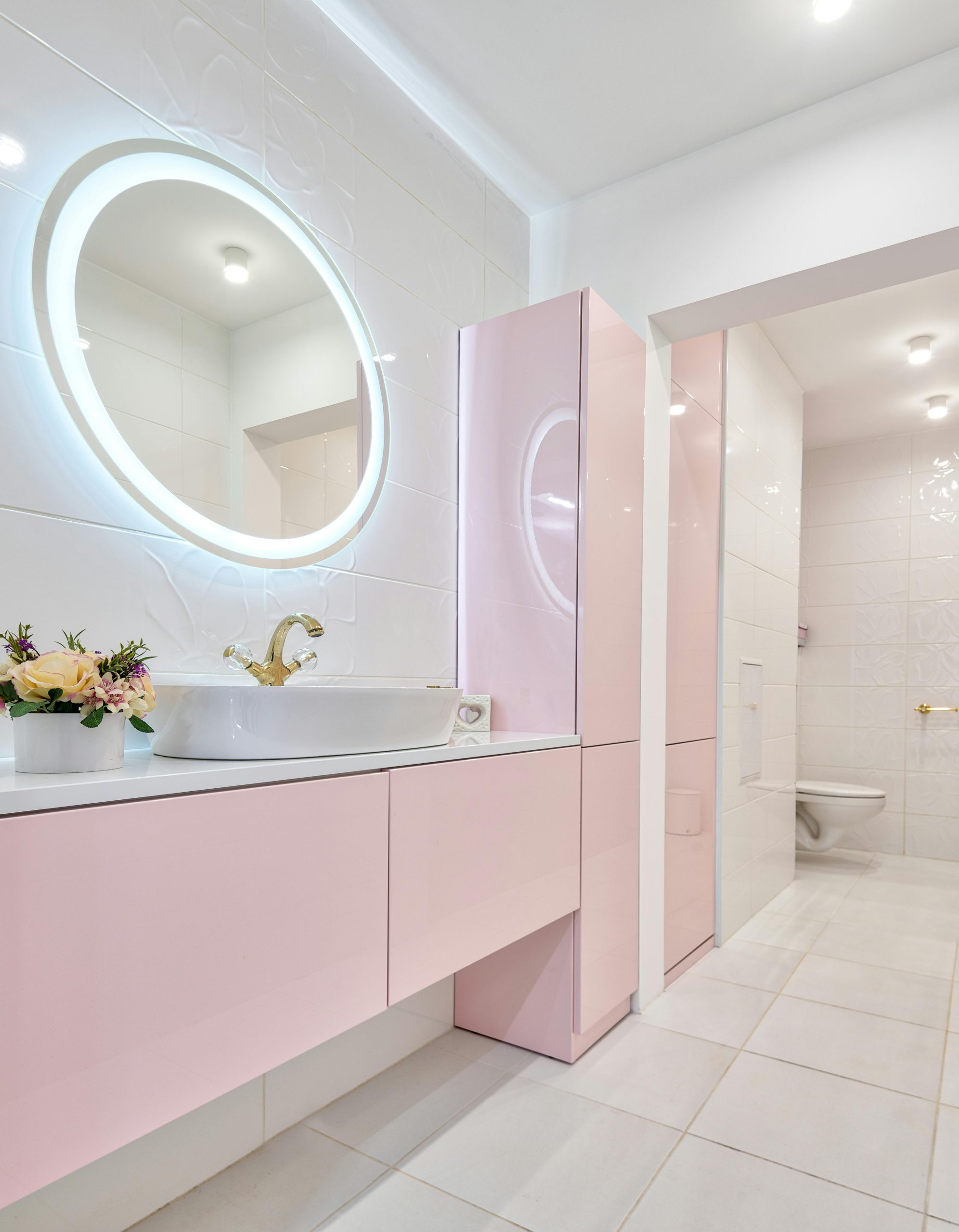 Modern restroom design with light pink cabinets and luminous mirror hanging above sink and decorative blooming flowers
