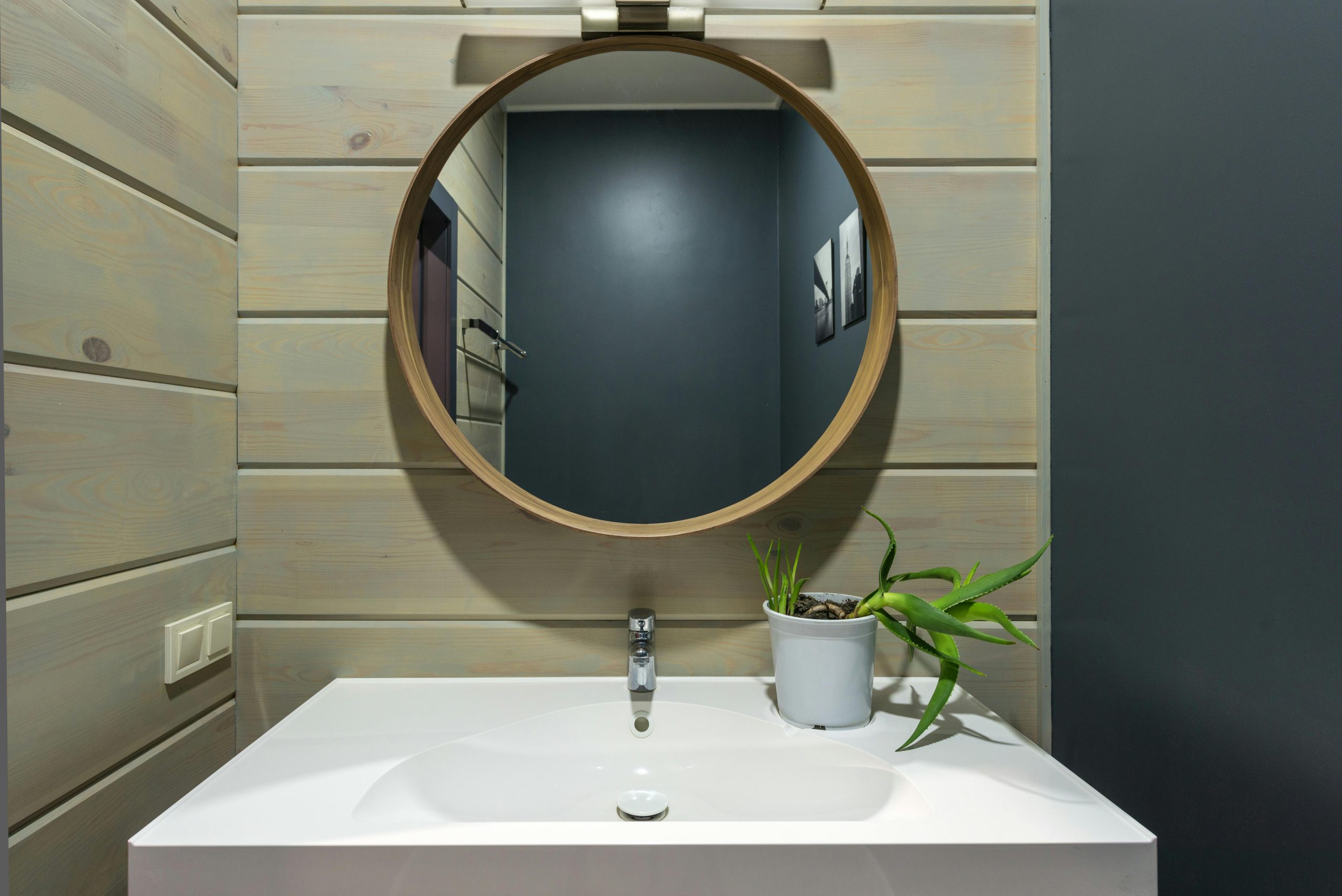 Round mirror hanging above sink with faucet and potted plant on wall made of panels in modern bathroom