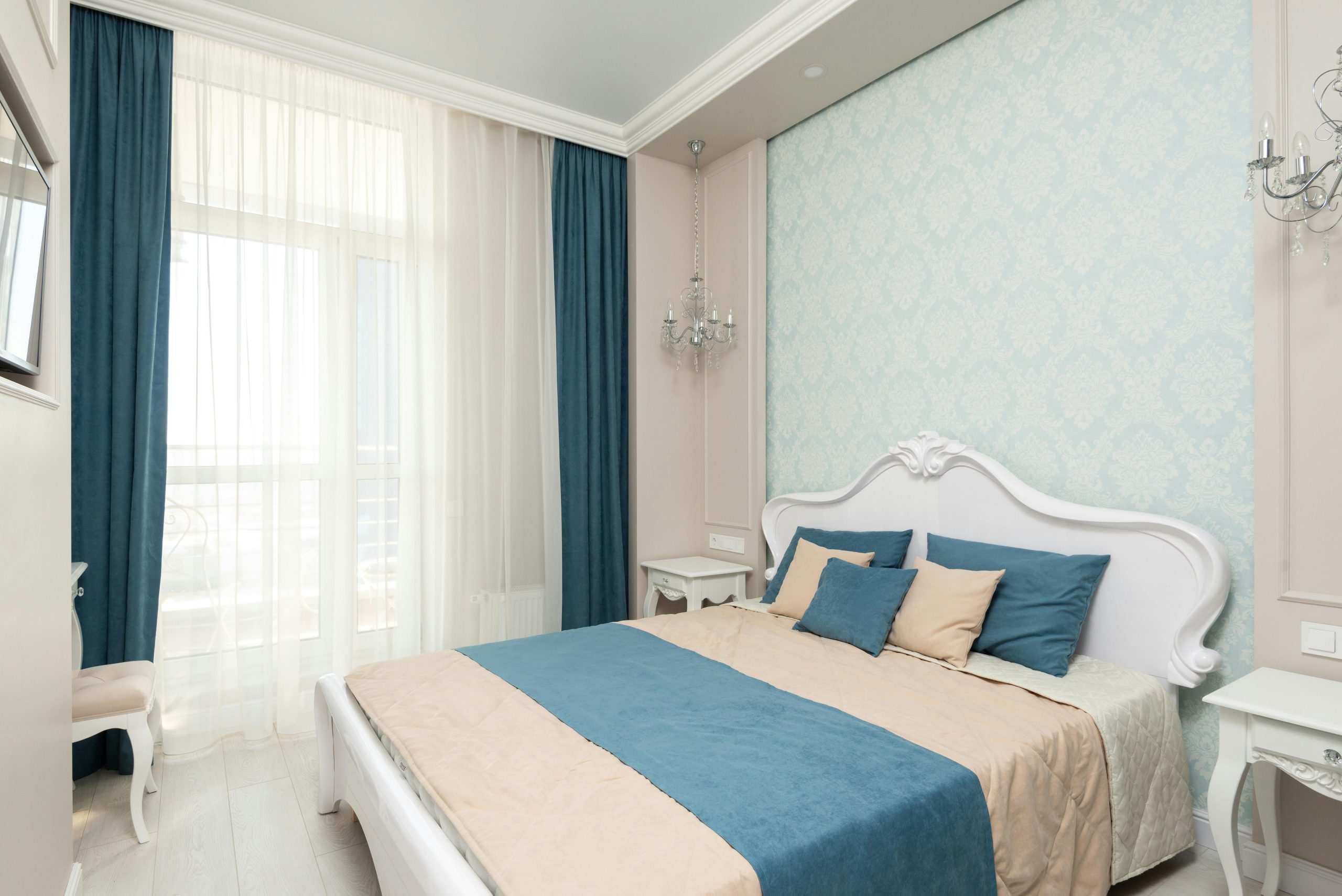 Soft bed with colorful bedclothes and cushions placed at wall with wallpaper near window with curtain in stylish spacious bedroom
