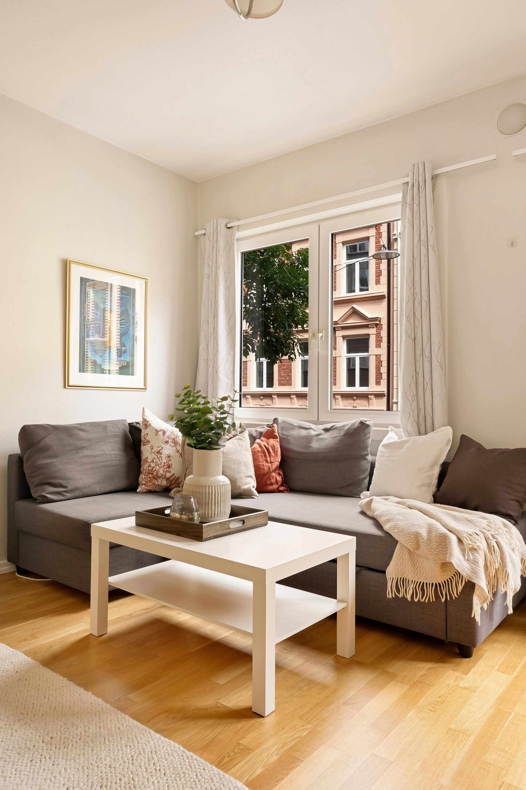 Stylish living room with a grey couch, wooden flooring, and a bright window view of neighboring building.
