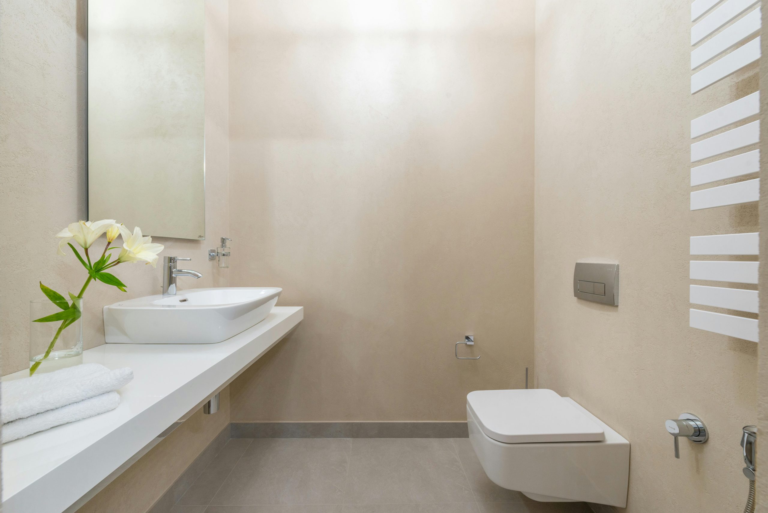 White ceramic toilet placed against wall with mirror and sink near blooming white flowers and towels in light modern bathroom