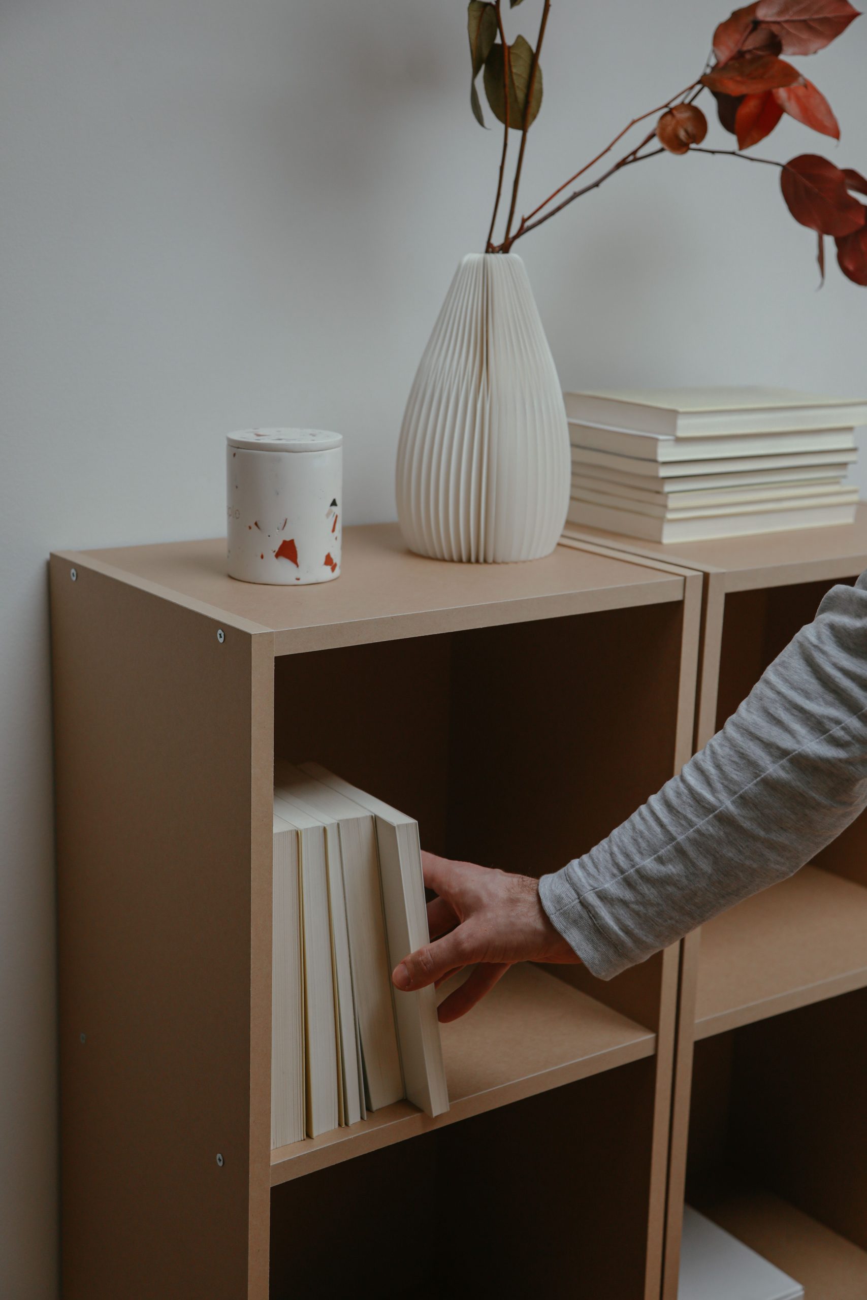 A hand reaches for a book on a minimalist bookshelf, next to decor and stacked books.
