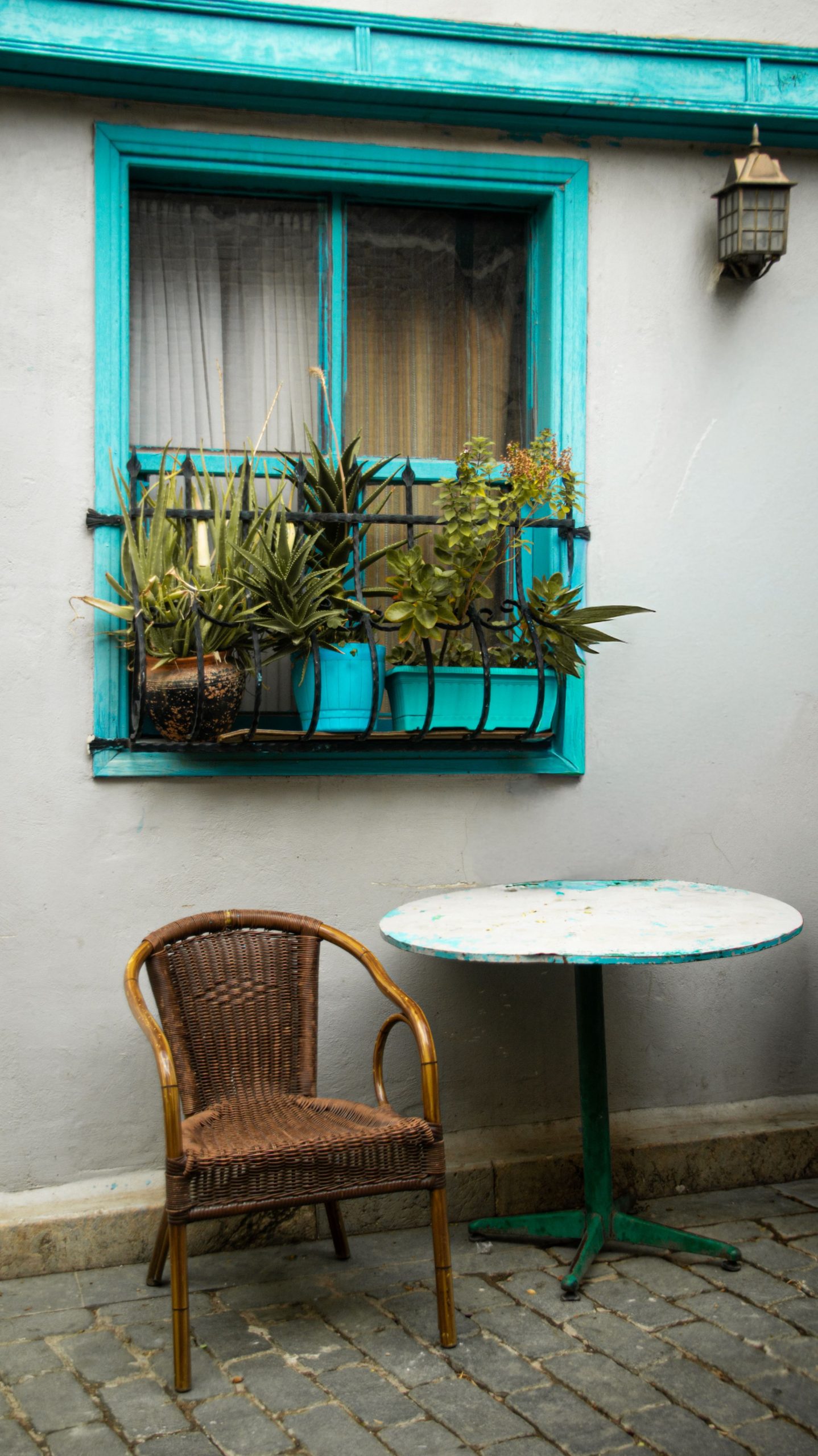 A quaint patio setting with a wicker chair, table, and window plants in a vibrant turquoise frame.