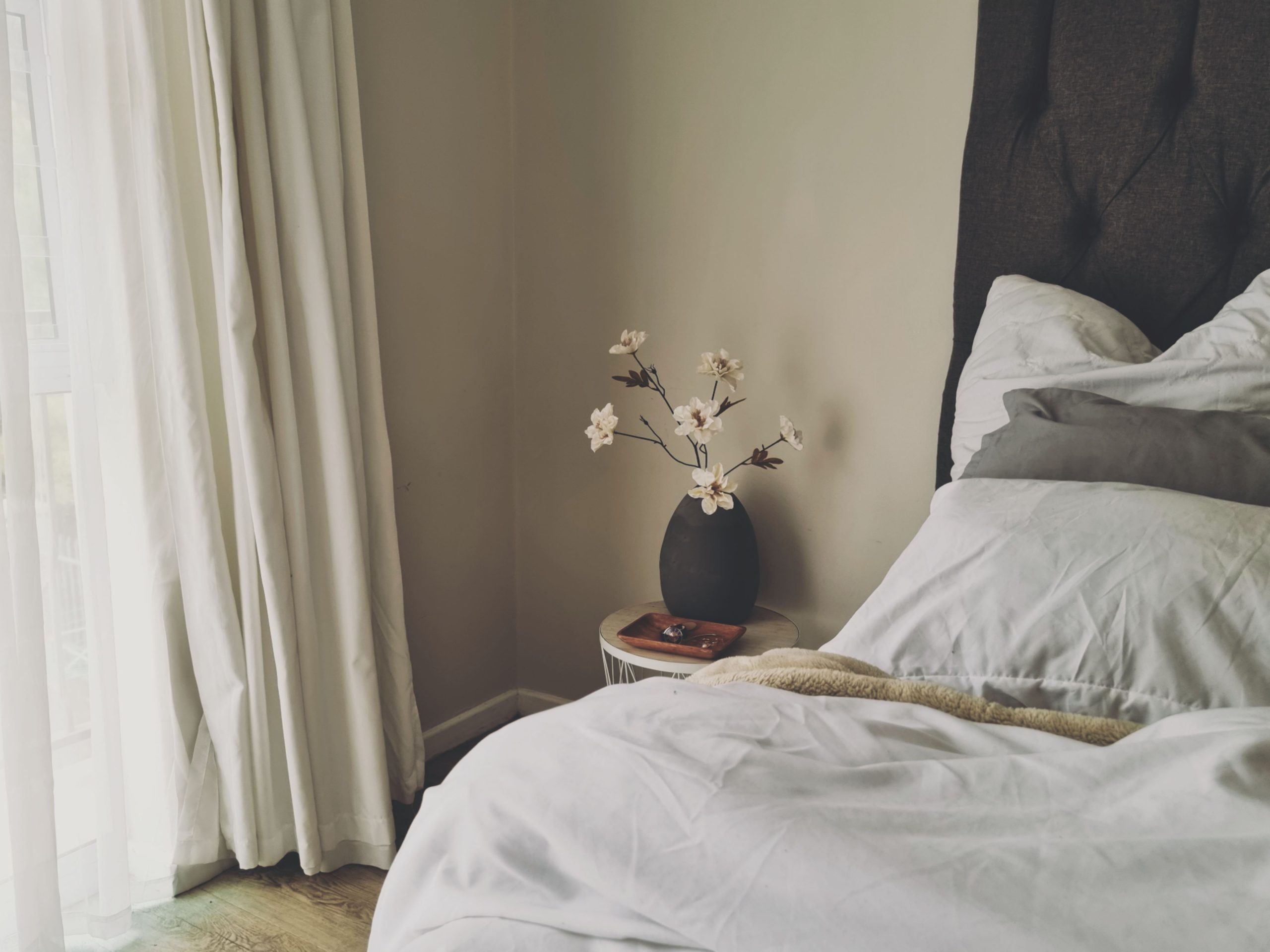A serene and minimalist bedroom interior featuring white flowers in a vase, natural light, and a cozy bedding setup.