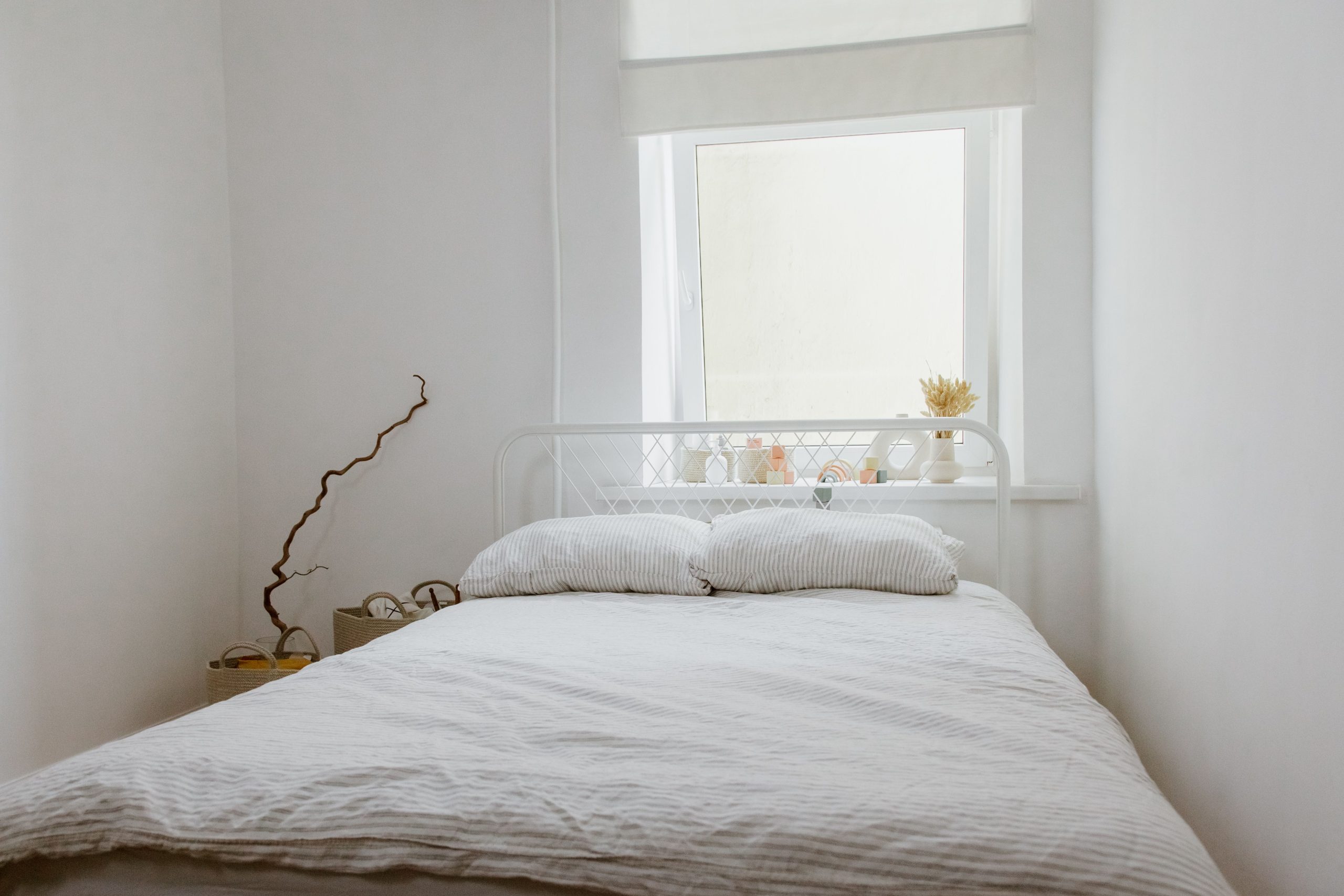 A serene white bedroom interior featuring a bed with pillows, natural light from a window, and minimalistic decor.