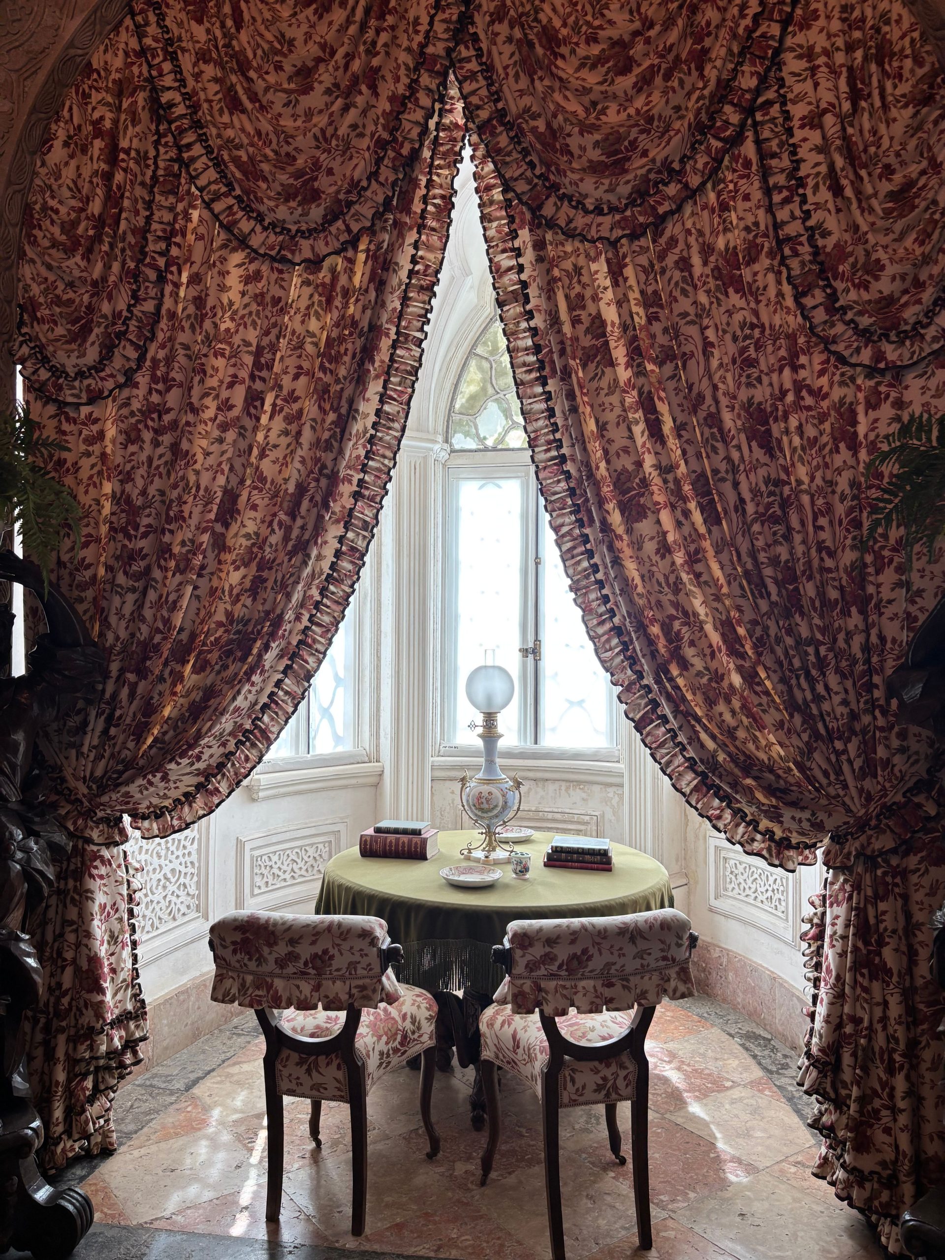 A vintage interior with floral drapes framing a Victorian-style table by a bright window.