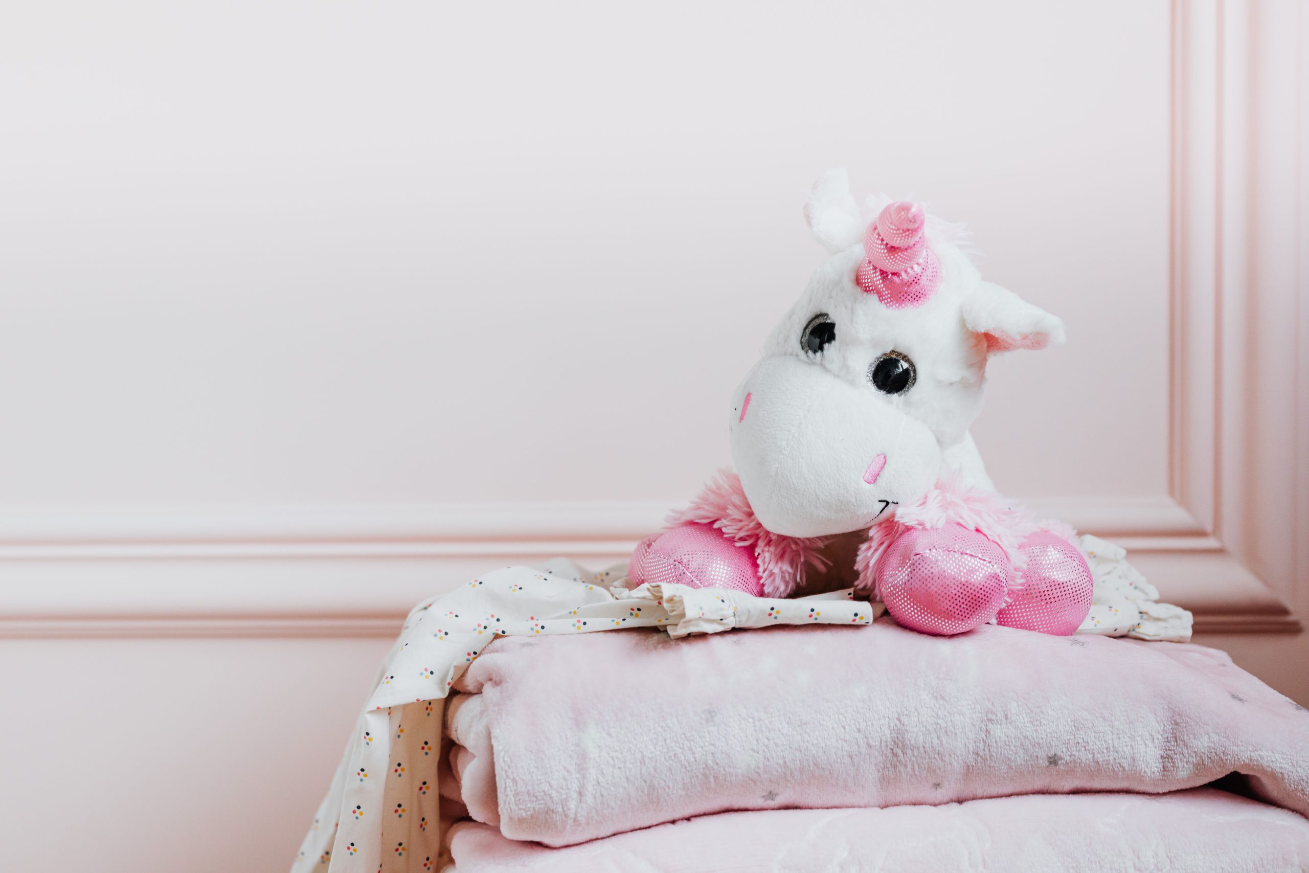 Adorable plush unicorn toy sitting on a stack of pink folded blankets in a cozy room.