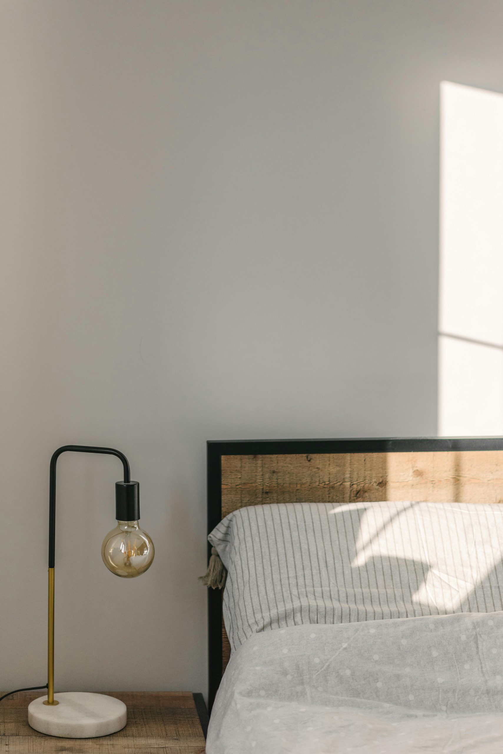 Bed with crumpled blanket near wooden bedside table with lamp and shadow on whitewashed wall at home
