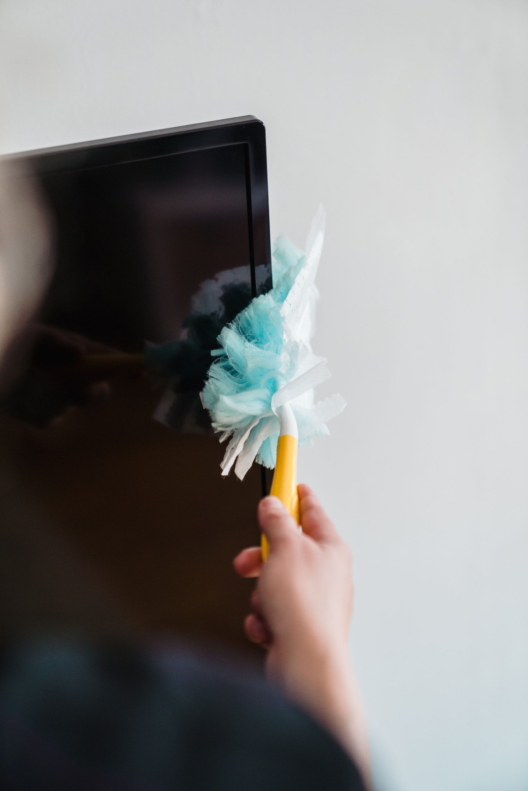 Close-up of a hand cleaning a TV screen with a blue feather duster indoors.