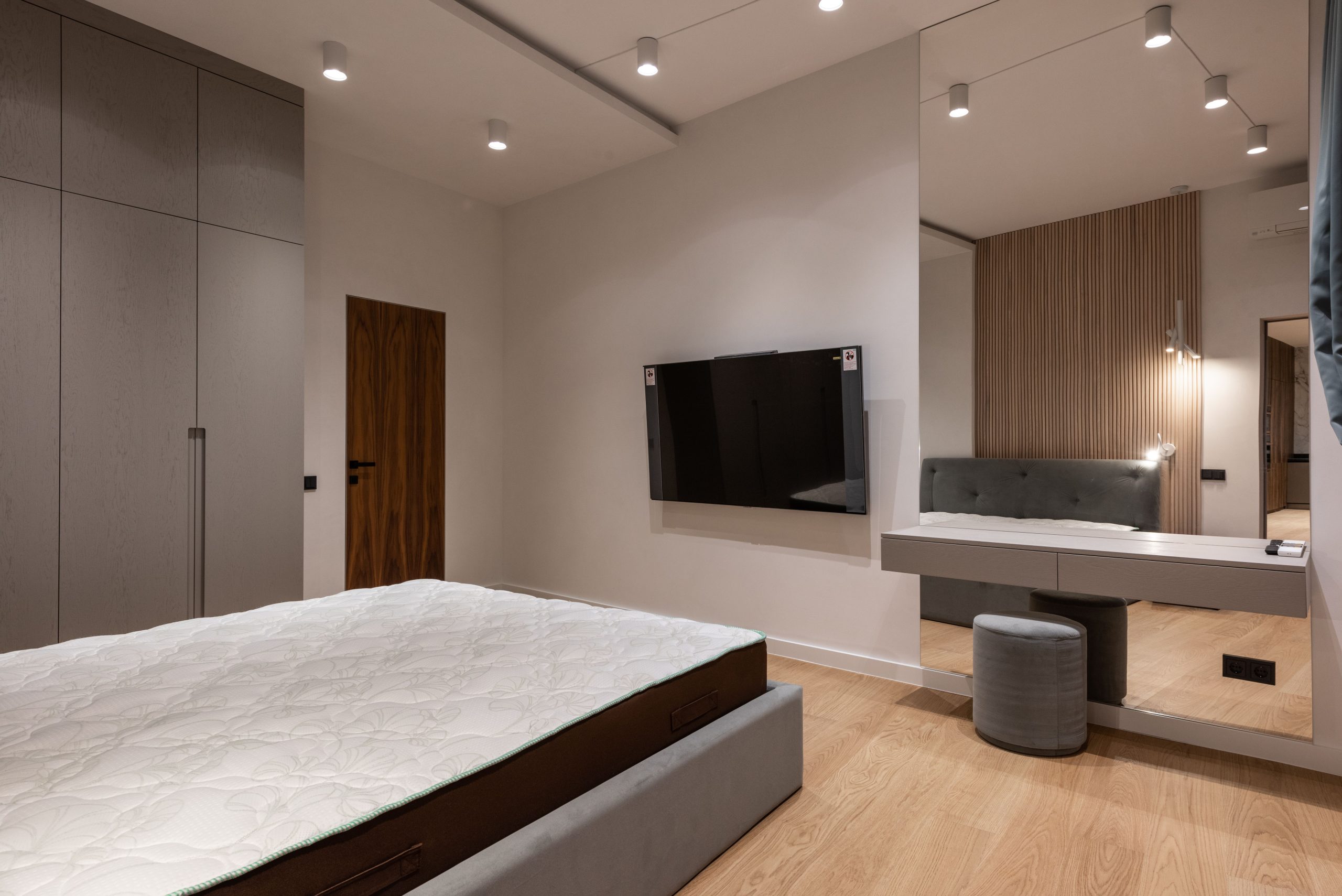 Comfortable bed and wardrobe in modern bedroom placed in front of TV hanging on wall near dressing table with big mirror
