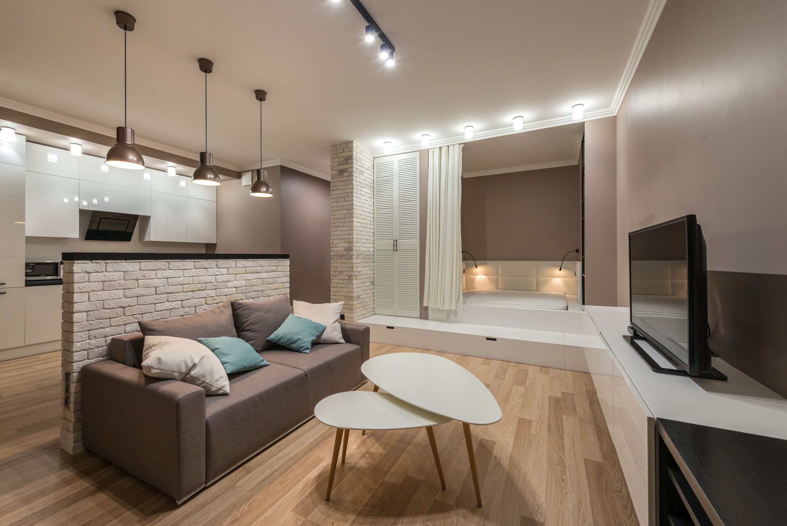 Interior of comfortable living room furnished with small sofa with cushions and white coffee table and TV and hanging lamps and illuminations on ceiling