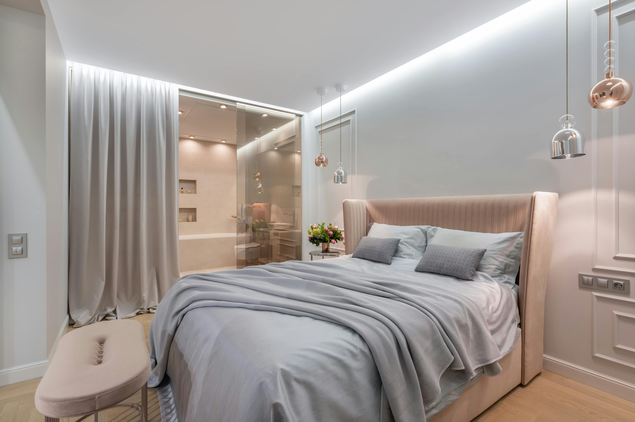 Interior of light modern home with bed with pillows and blanket next to pouf and table under lamps near glass door with curtain to washroom