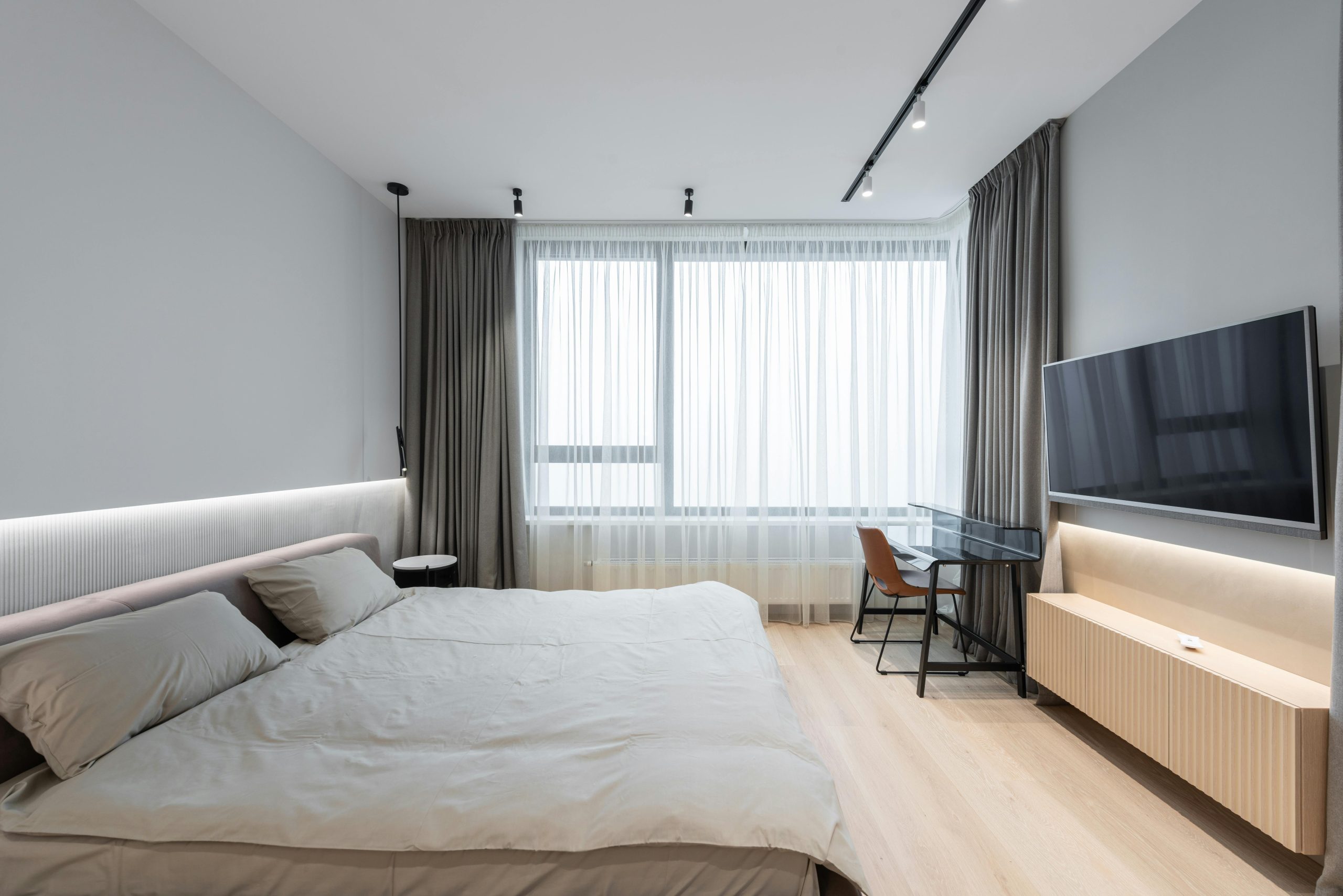 Interior of modern bedroom with bed next to TV on wall with curtains on window and table with chair