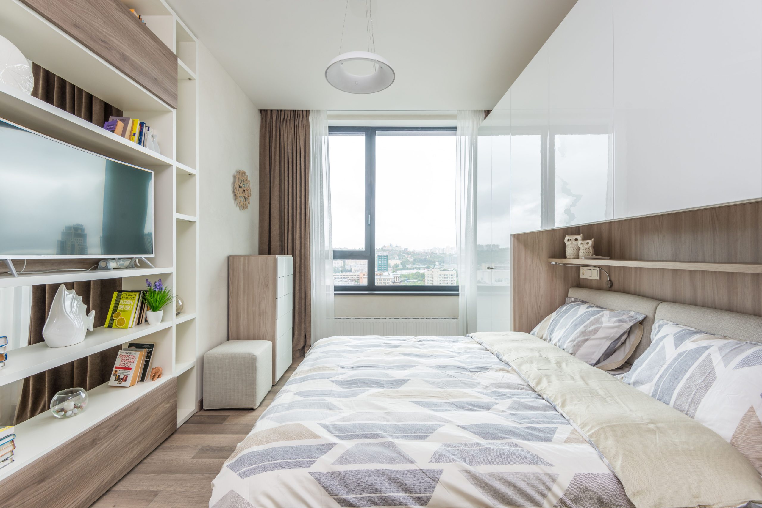Interior of modern bedroom with comfortable double bed with bedside wardrobe near window and shelves with decor and TV