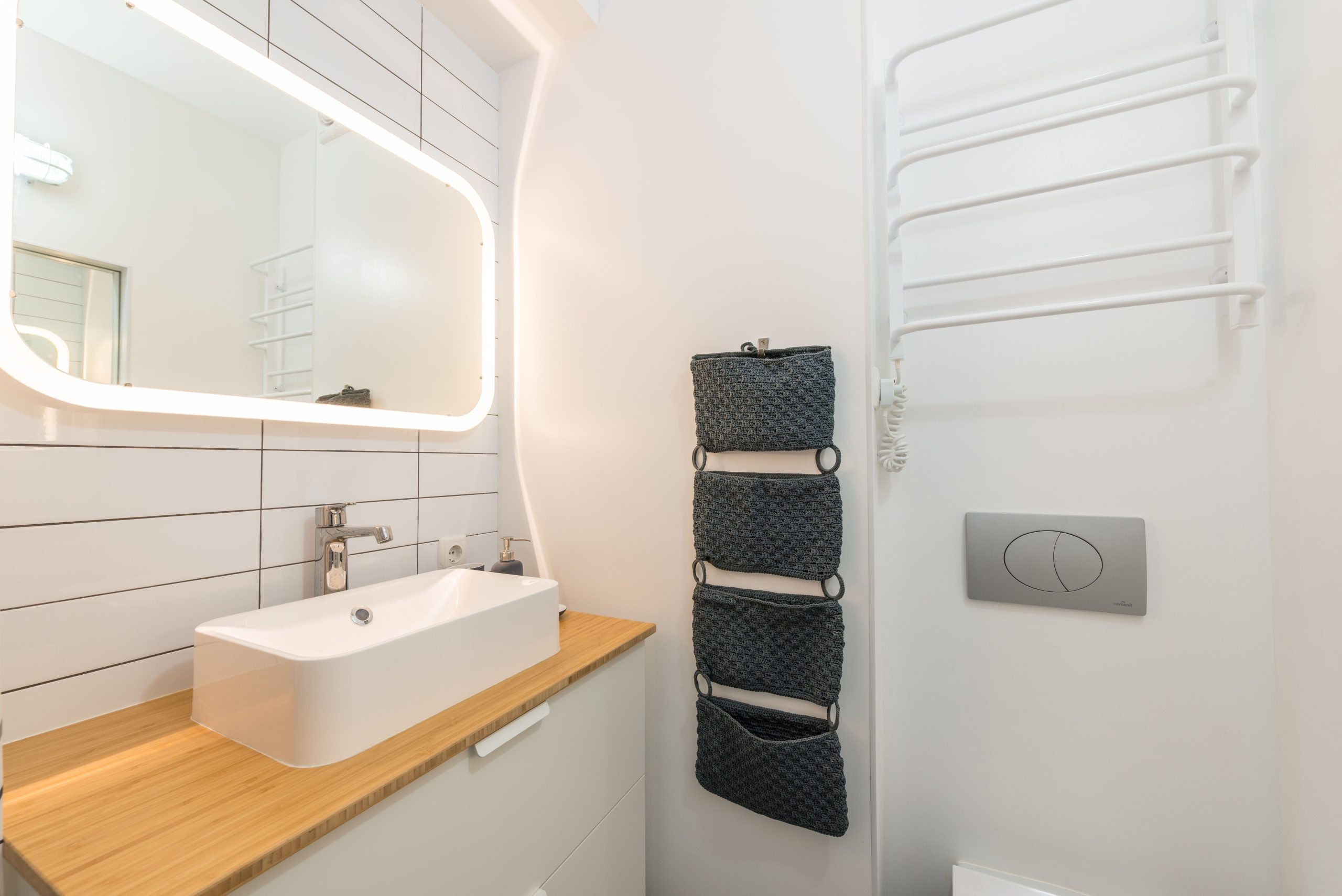 Interior of small modern bathroom with ceramic sink placed on white cabinet with wooden top under illuminated mirror