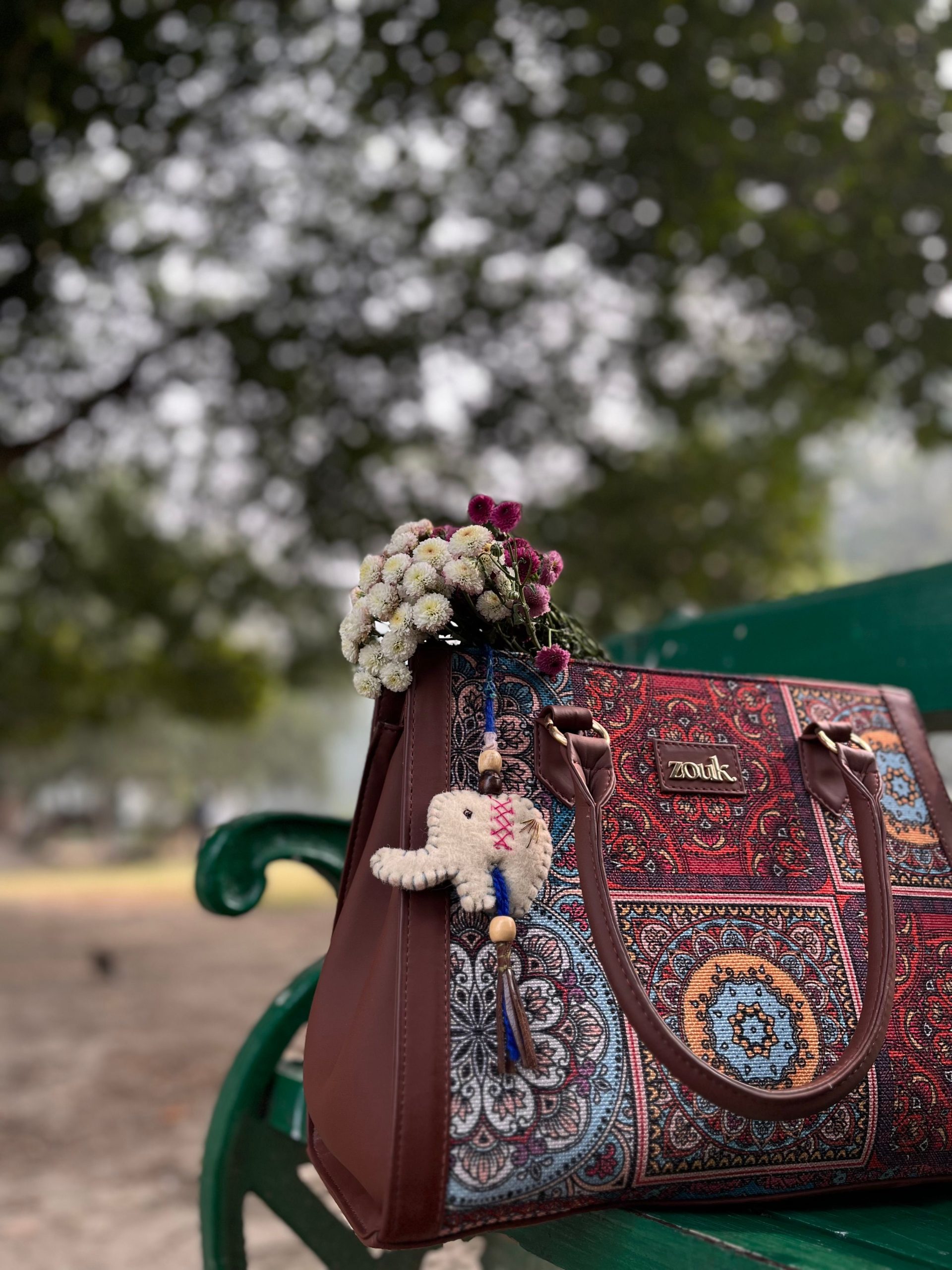 Ornate patterned handbag with floral accessory on a park bench, lush outdoors setting.