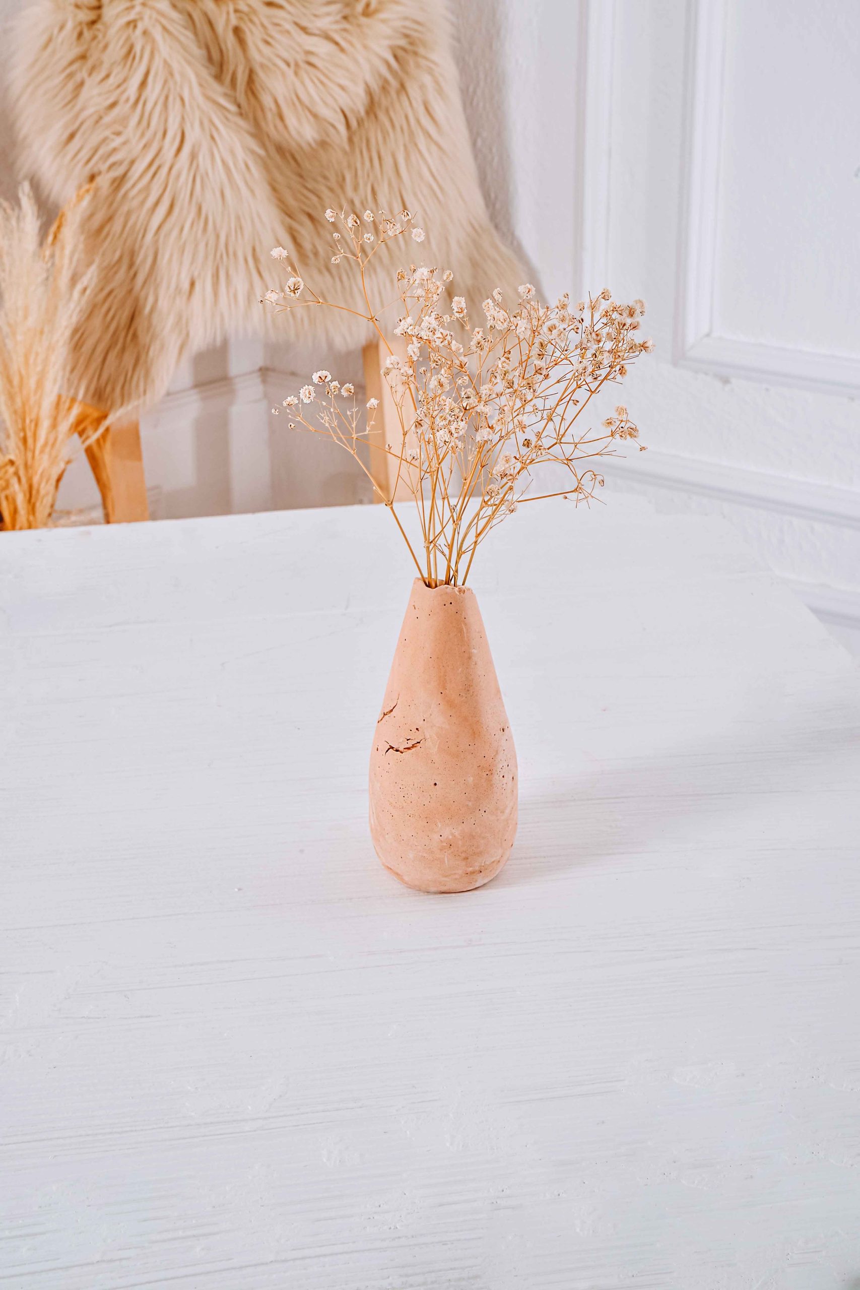 Stylish minimalist setup featuring a clay vase with dried flowers on a white table in a cozy interior.