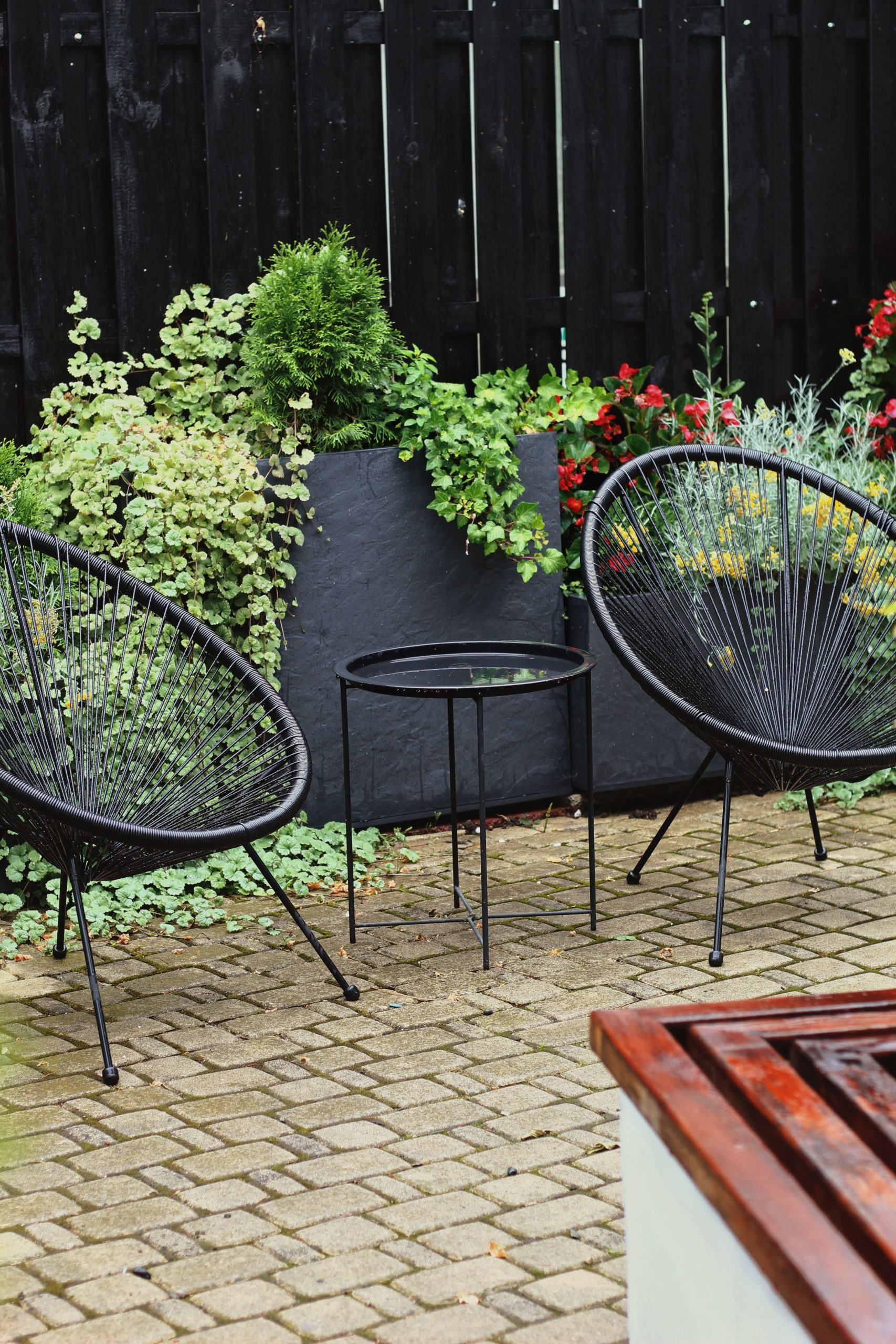 Stylish outdoor patio setup with black wire chairs, small table, and lush greenery.